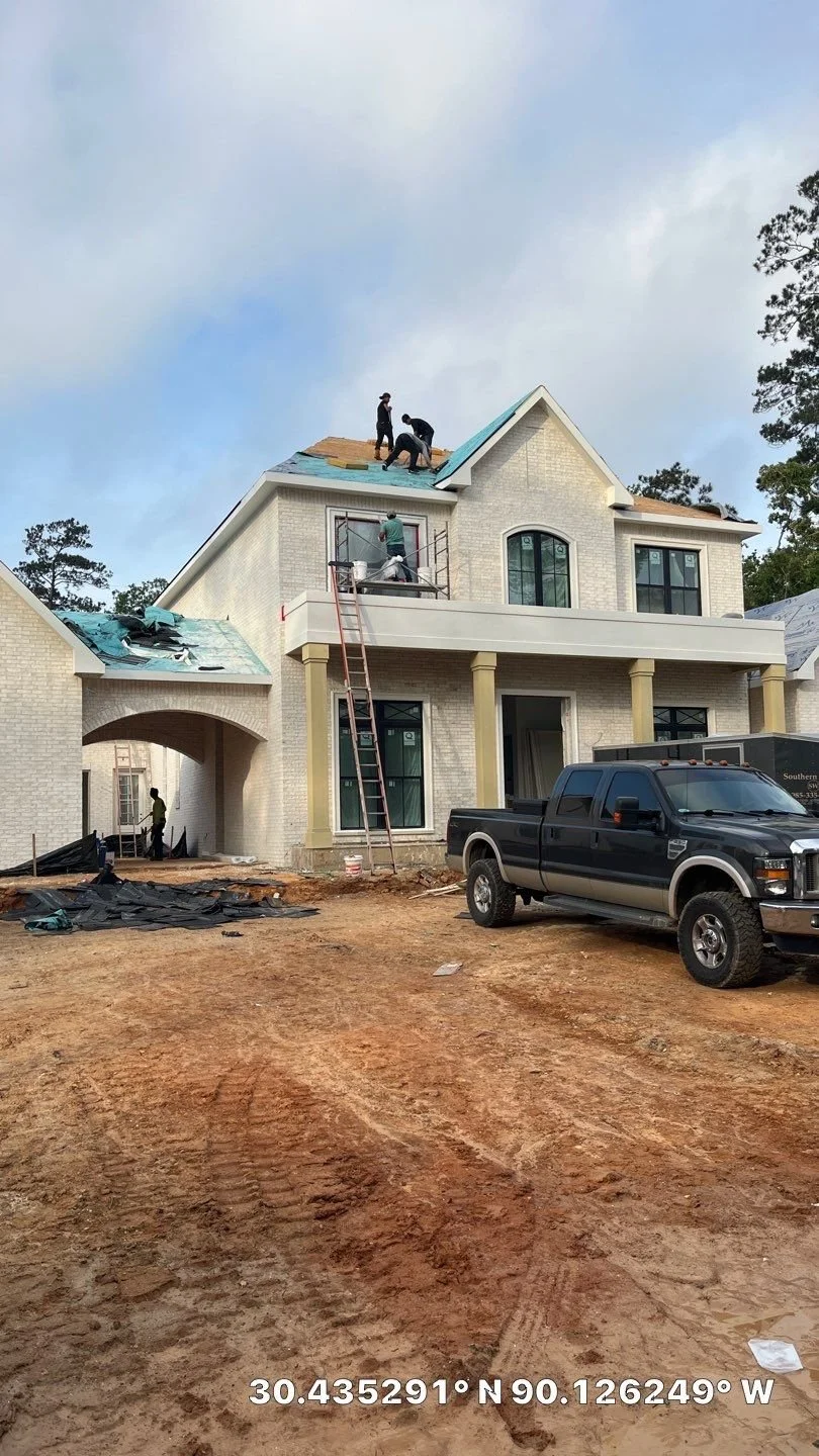 Construction workers installing a roof on a large two-story house with a brick exterior, columns, and large windows, parked truck in front, and dirt ground.
