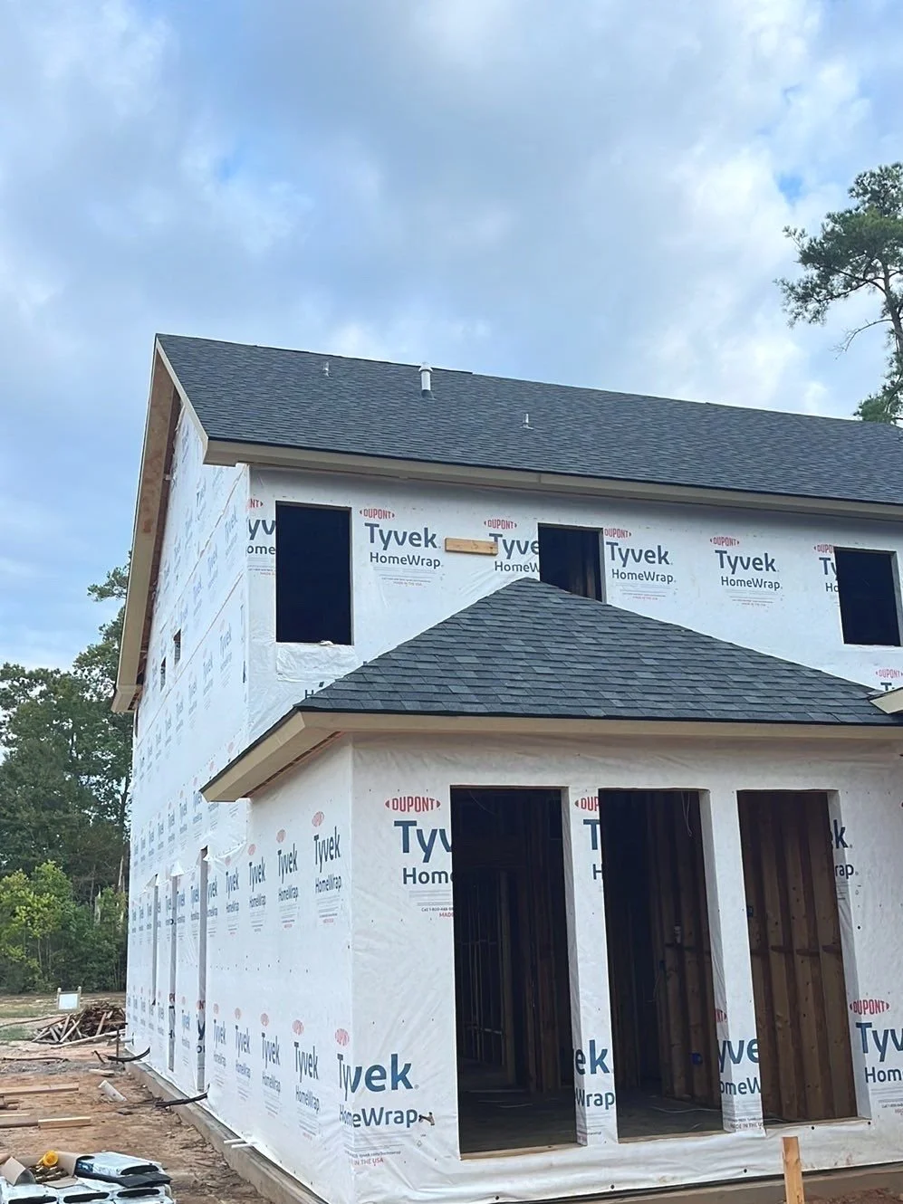 A house under construction wrapped with Tyvek HomeWrap, with some windows and door openings visible, and a completed roof with asphalt shingles.