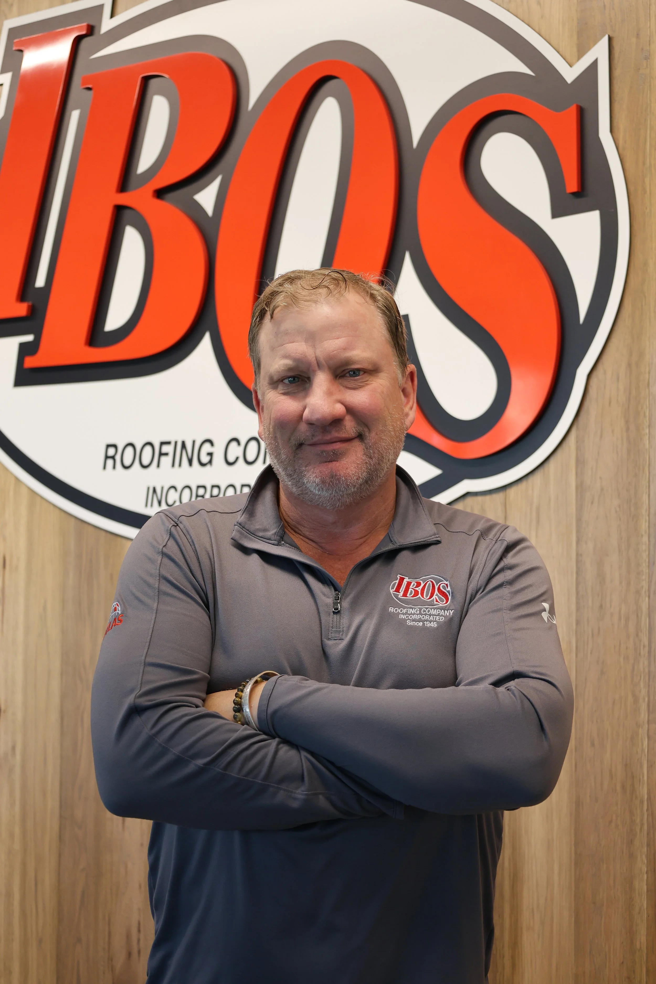 A man with crossed arms wearing a navy blue polo shirt with a company logo stands in front of a sign that reads 'IBOS' and 'INC'.