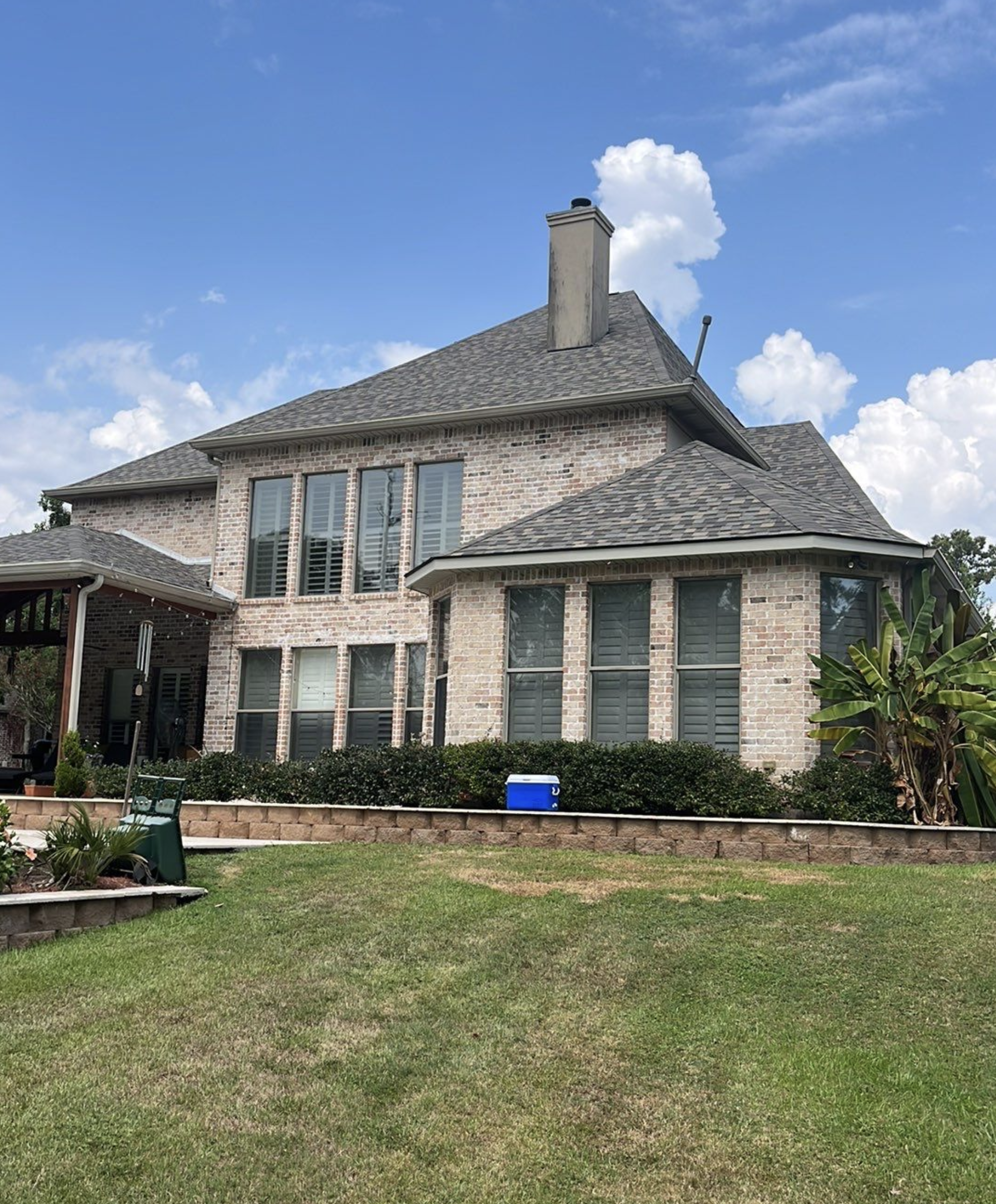 Back view of a two-story brick house with large windows, a chimney, and a well-maintained lawn under a partly cloudy sky.