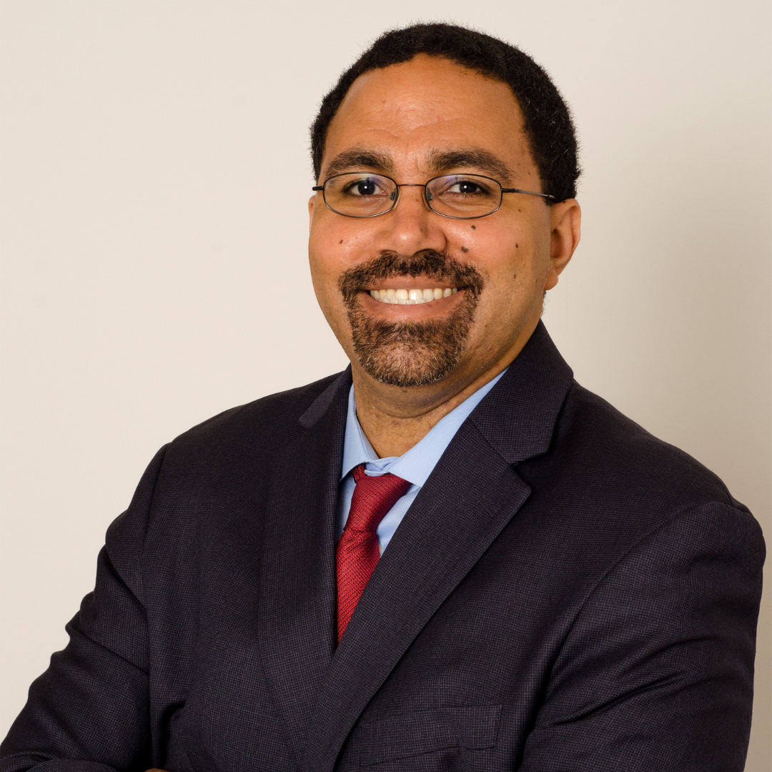 A man with glasses, a goatee, and short curly hair, wearing a dark suit, light blue shirt, and red tie, smiling at the camera against a plain white background.