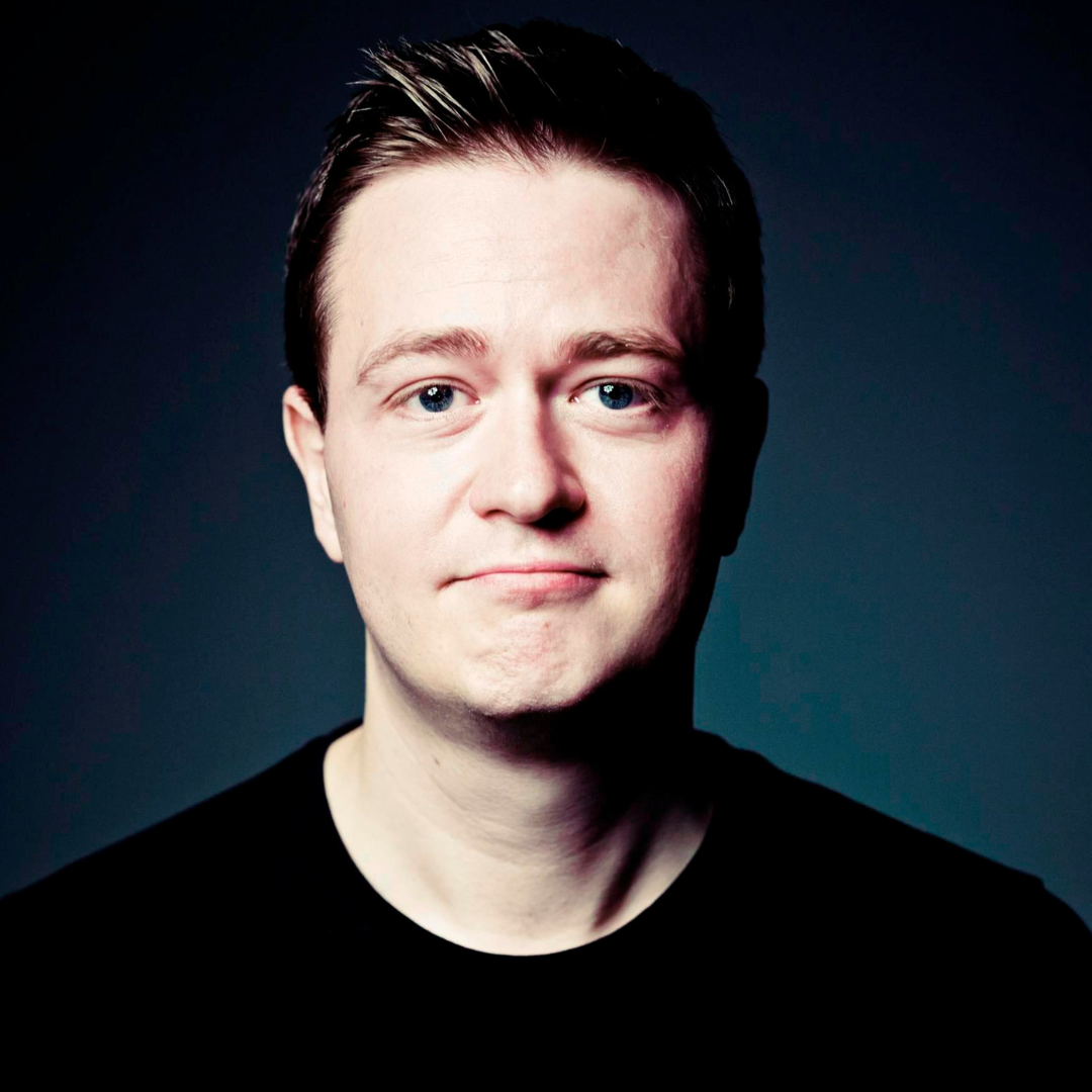 Portrait of a young man with light skin, short brown hair, blue eyes, wearing a black shirt, against a dark background.