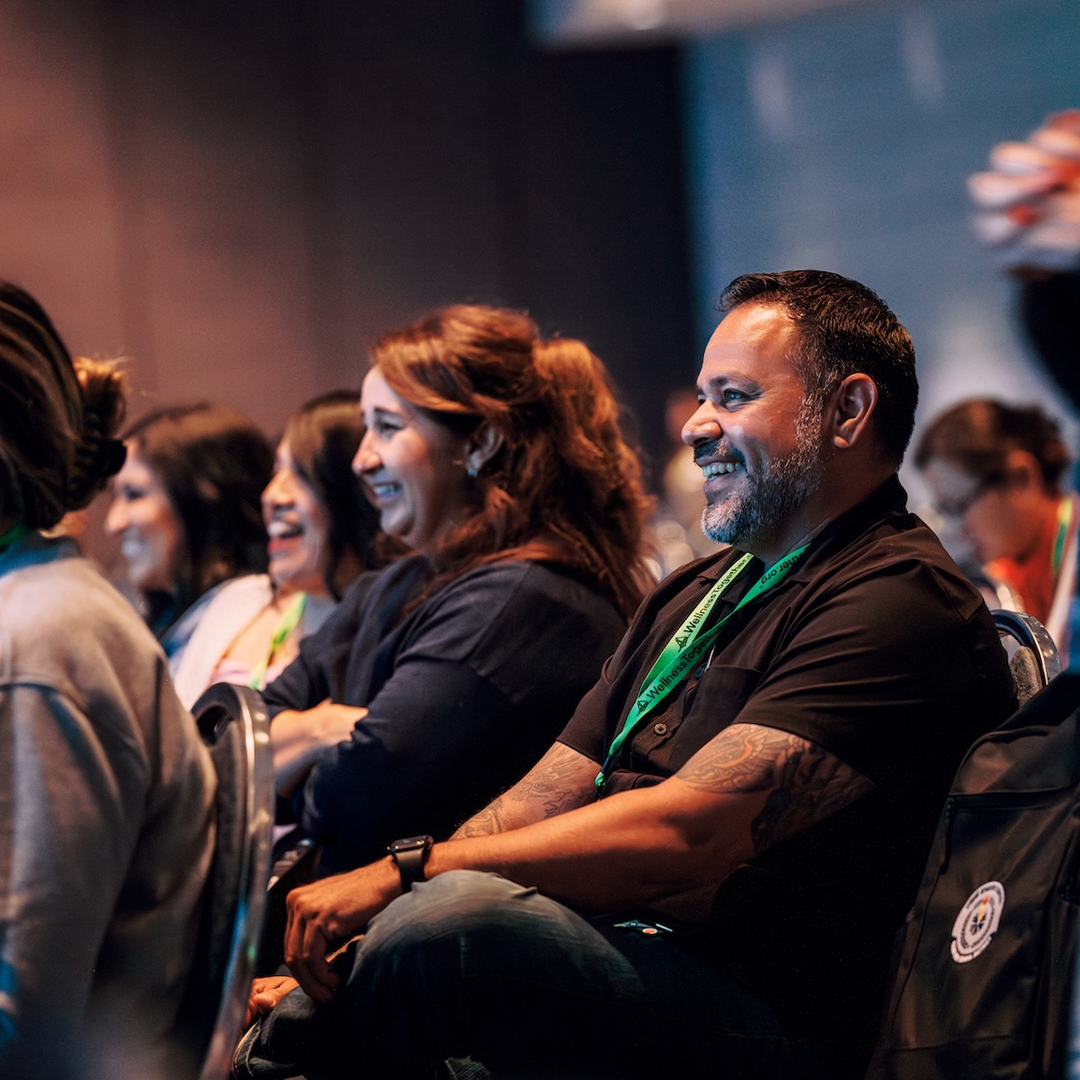 People seated and smiling at a conference or seminar, with a focus on a man with tattoos and a beard in the foreground.