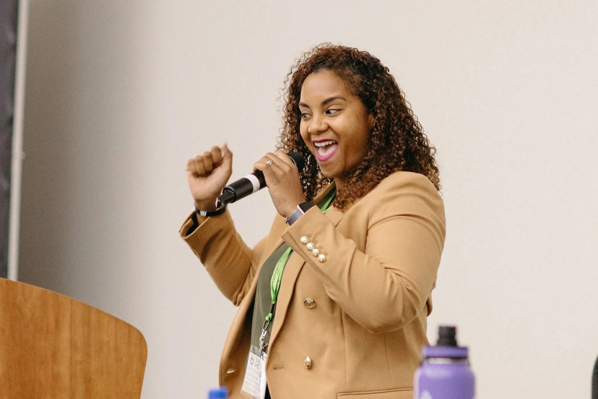 A woman speaking at a conference, wearing a burgundy dress, with a conference badge around her neck, standing in front of a podium with a laptop, at the 8th Annual Wellness Together Conference.