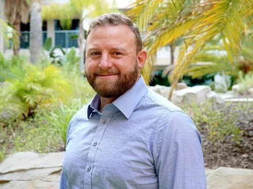 A man with a beard smiling outdoors with plants and palm trees in the background.