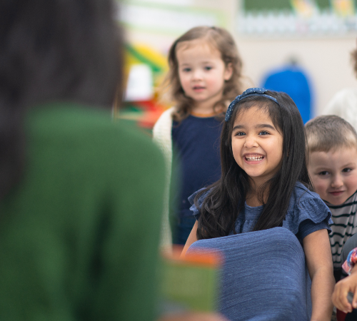 Smiling young girl in a classroom with other children watching an adult.