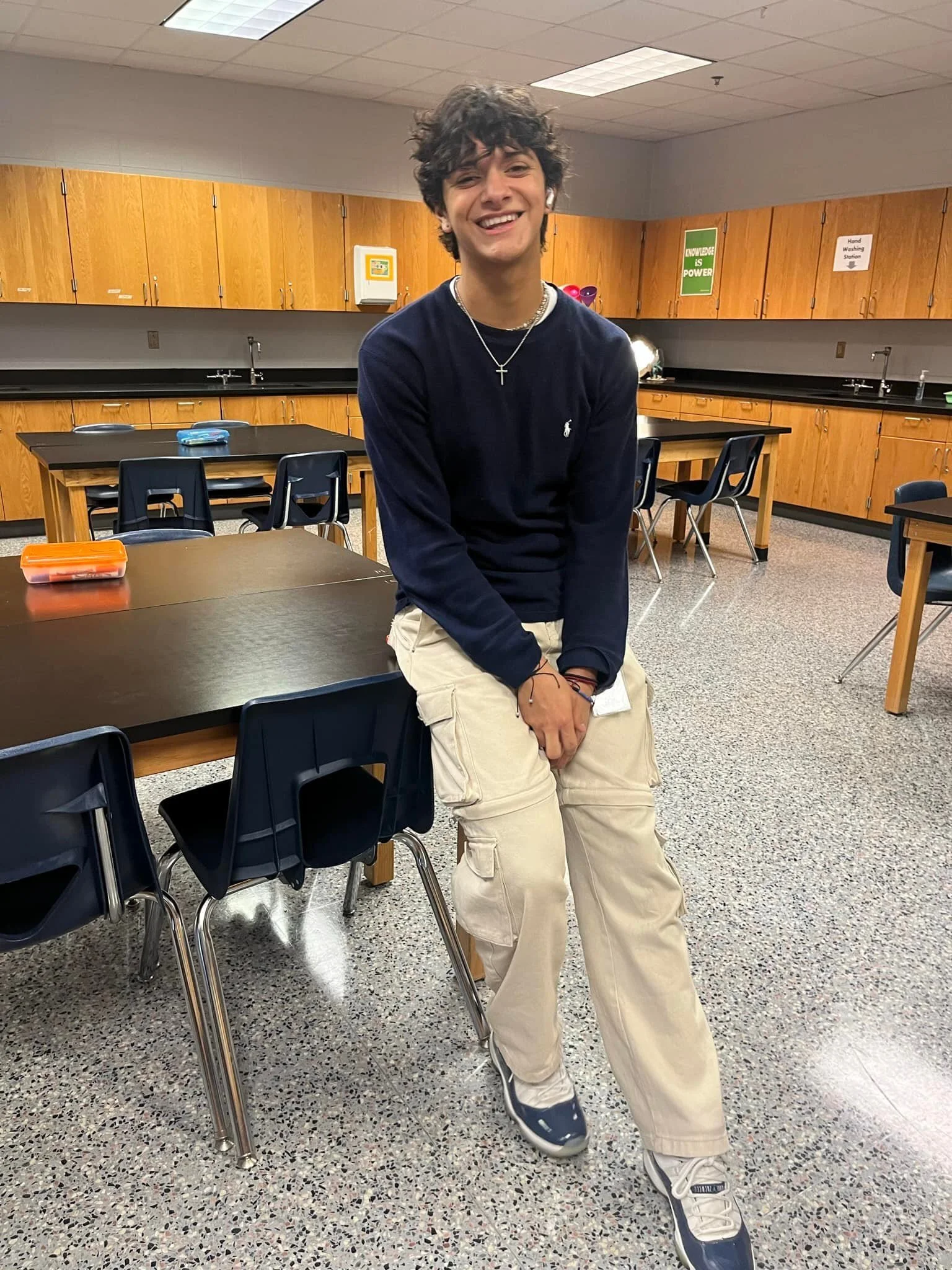 A young man with curly dark hair, wearing a navy blue sweater, beige cargo pants, and sneakers, sitting on a chair in a classroom or cafeteria with wooden cabinets, black countertops, and several black chairs and tables.