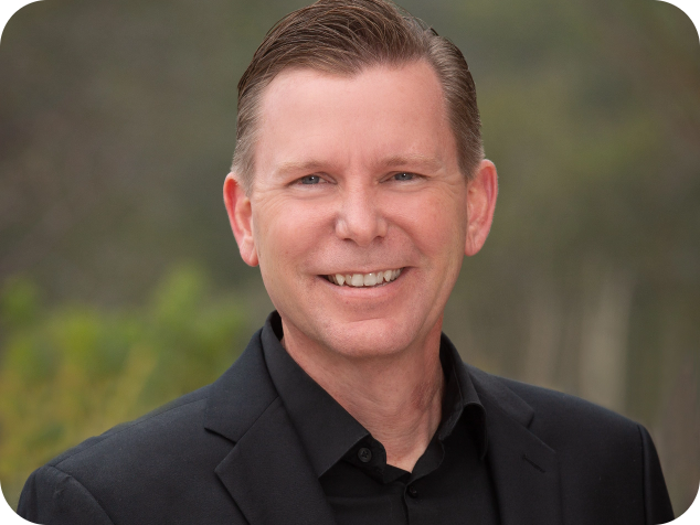 A smiling man in a black suit jacket and shirt, outdoors with blurred greenery in the background.