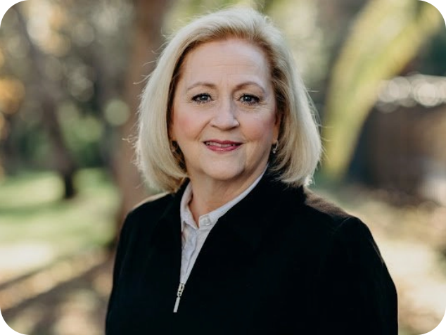 A middle-aged woman with blonde hair smiling outdoors, wearing a black jacket and white shirt.