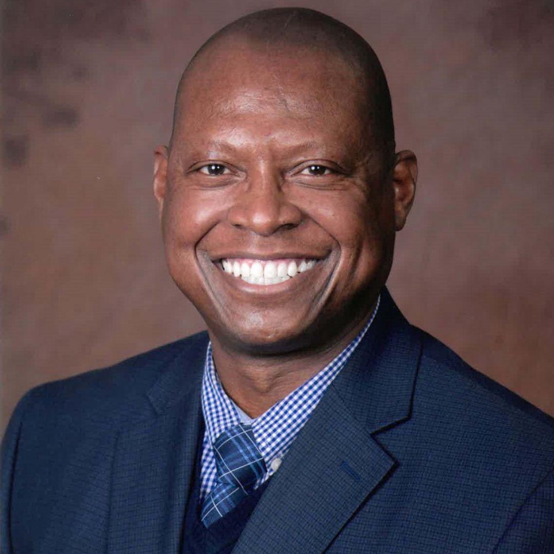 Professional headshot of a smiling African American man wearing a blue checkered shirt, a blue tie, and a dark blue blazer with a brown background.