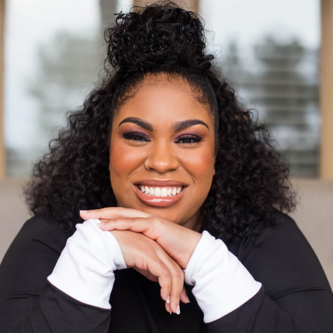 A smiling woman with curly black hair styled in a top bun, wearing makeup, a nose ring, and a black top with white sleeves, sitting indoors with a blurred window in the background.