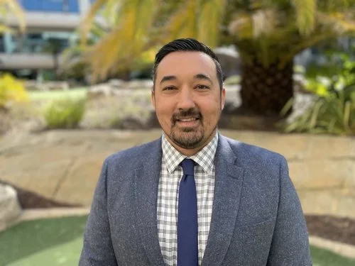 A man in a suit standing outdoors with trees and greenery in the background.