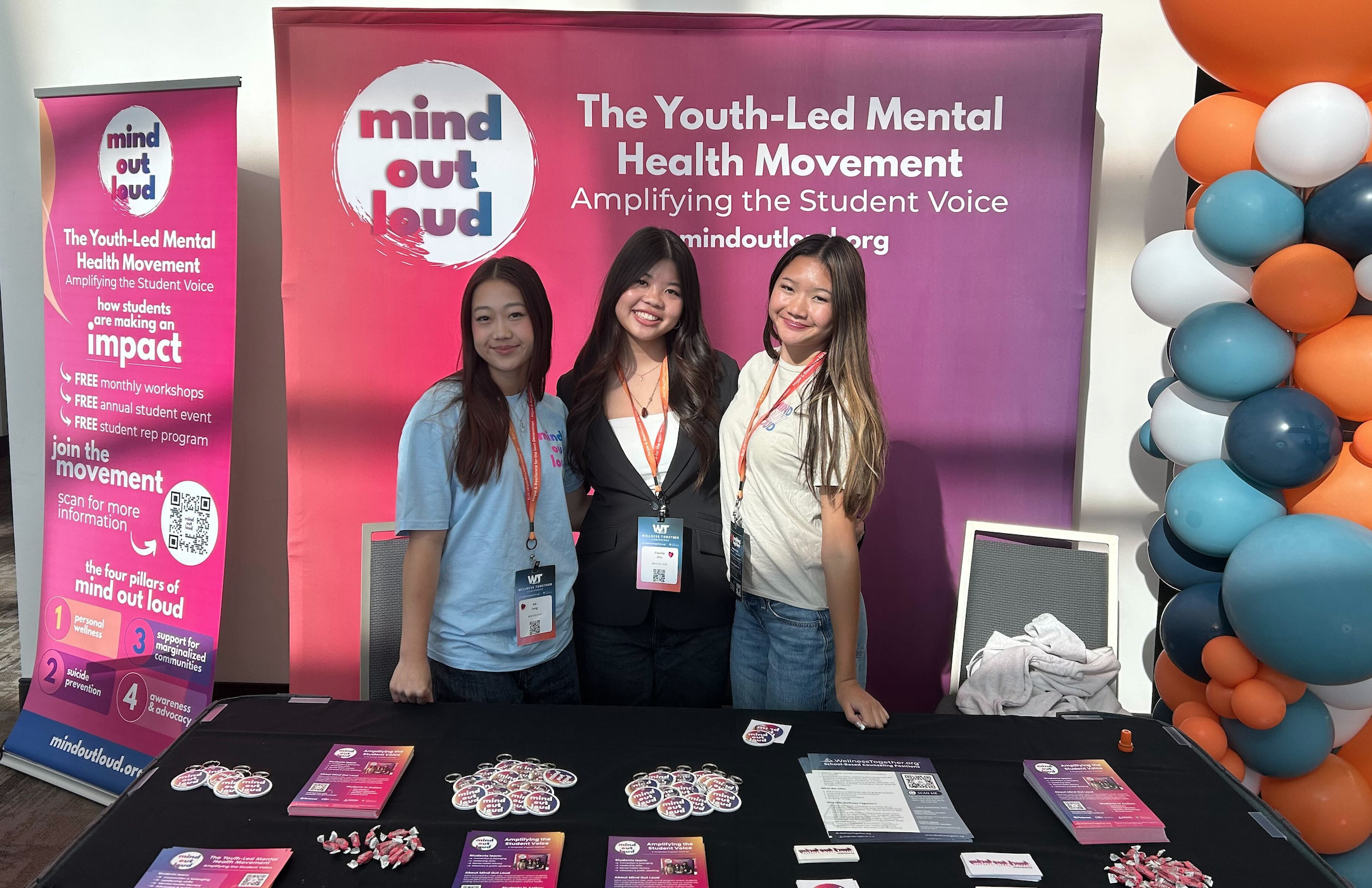 Three young women standing behind a table at a mental health event, smiling, with a pink and purple backdrop that reads 'mind out loud' and information about youth-led mental health movement, plus a pink banner on the left with event details and a QR code, and colorful balloons on the right.