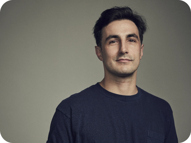 A man with dark hair and a slight smile, wearing a navy blue sweater, standing against a plain gray background.