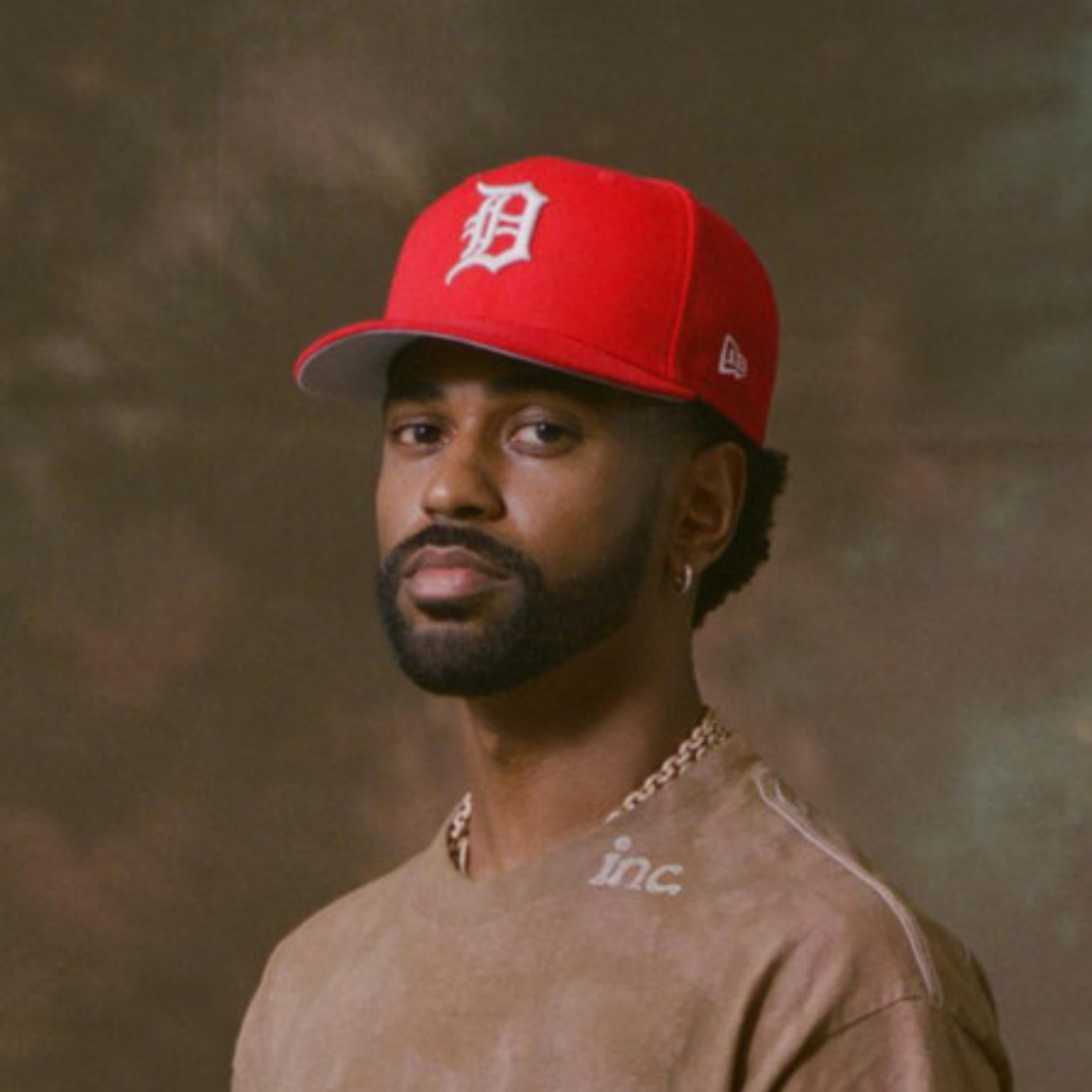 A young man with a beard and mustache wearing a red baseball cap with a white 'D' logo and a beige T-shirt, standing against a neutral background.