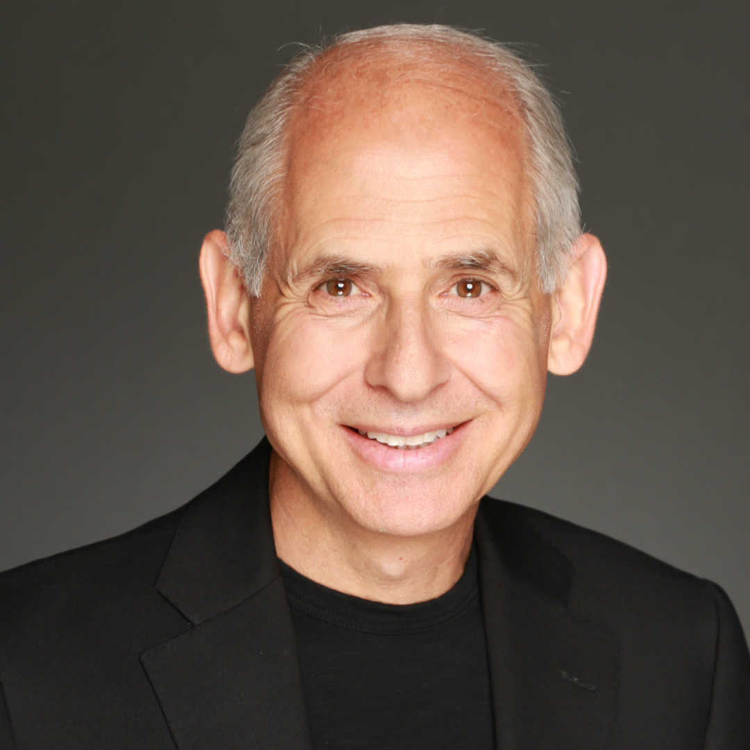 A smiling older man with short gray hair, wearing a black blazer and a black shirt, against a dark gray background.