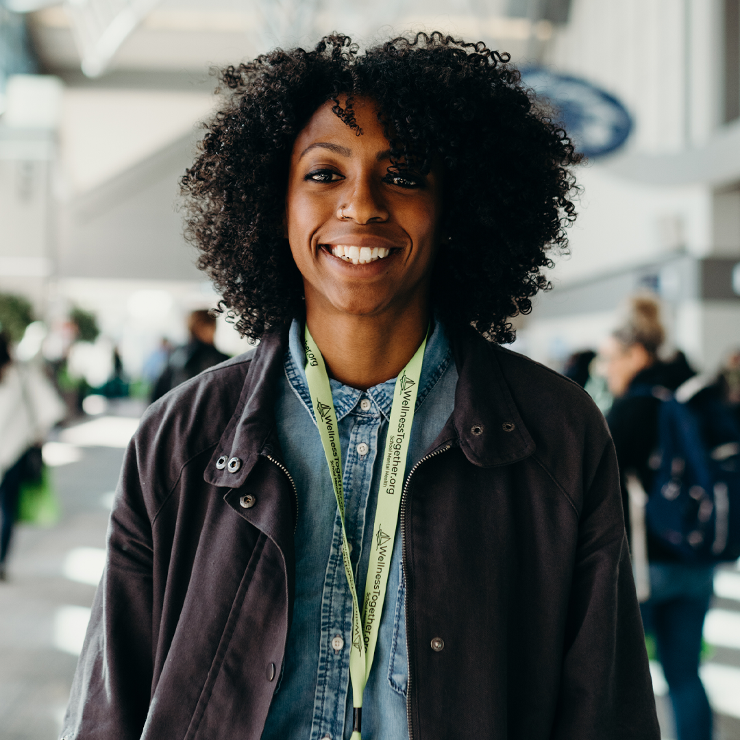 A smiling woman with curly black hair wearing a denim shirt, black jacket, and a lanyard, standing in a busy indoor setting.