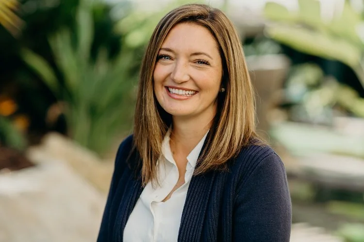 A smiling woman with shoulder-length brown hair wearing a white blouse and navy blazer outdoors with green foliage in the background.