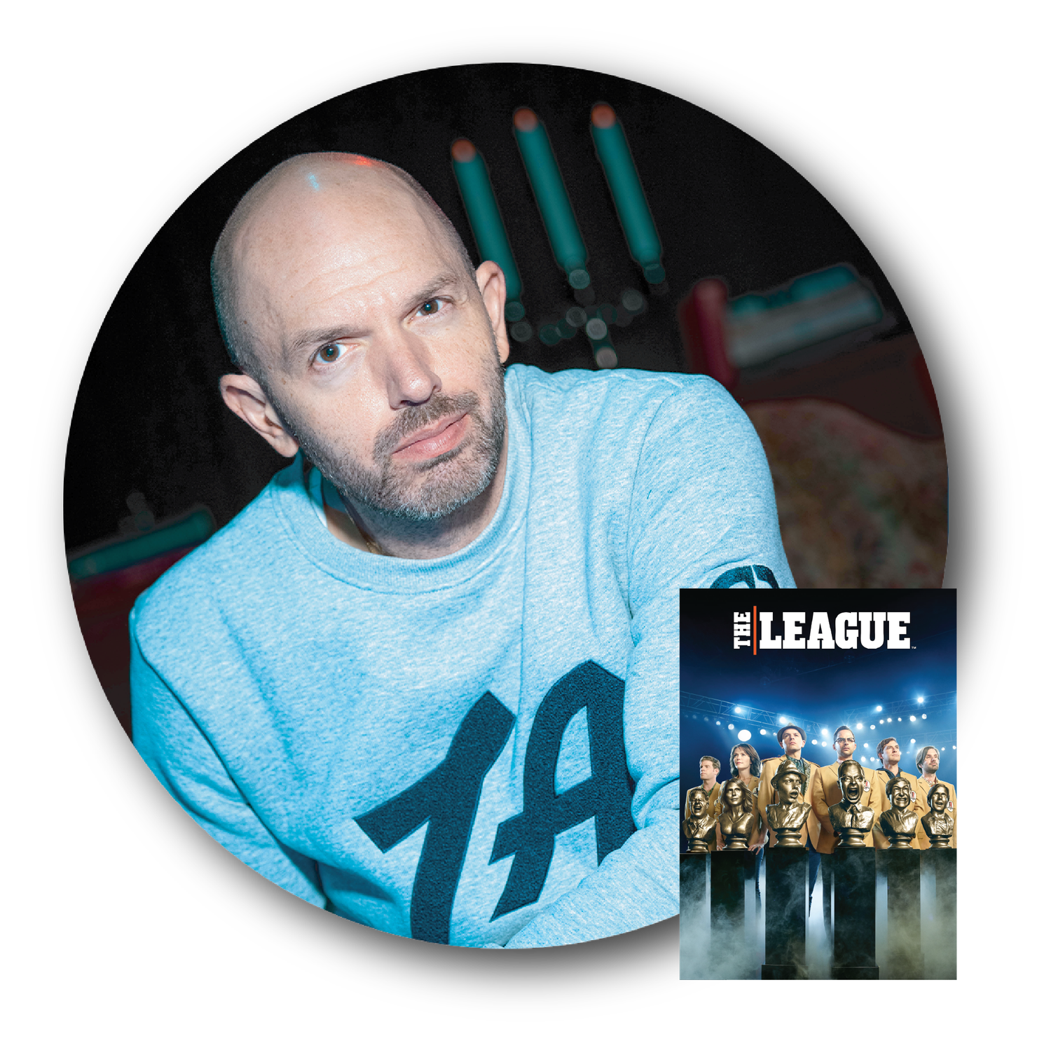 A man with glasses and blond hair wearing a light blue shirt, sitting at a desk with framed artwork behind him.