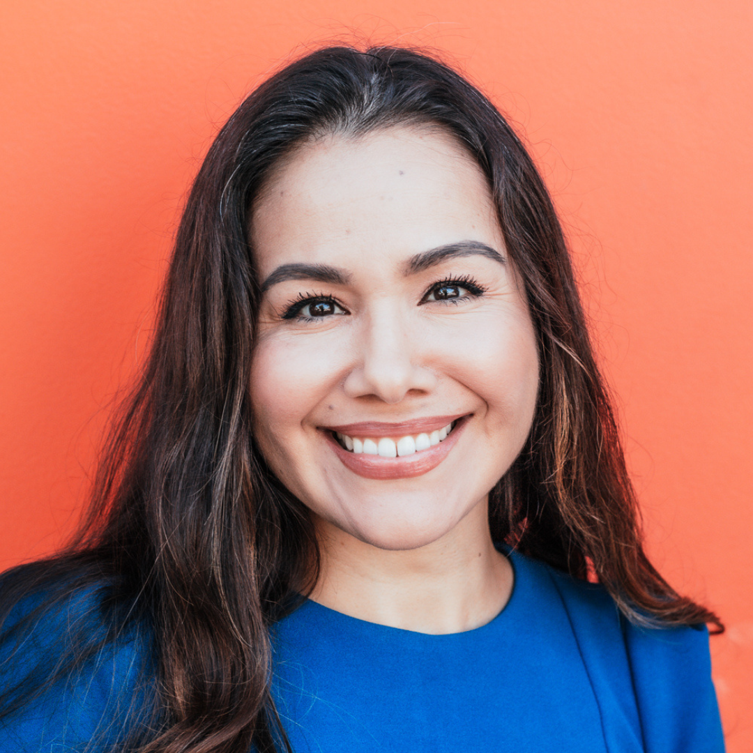A woman with long dark hair smiling against an orange background, wearing a blue top.