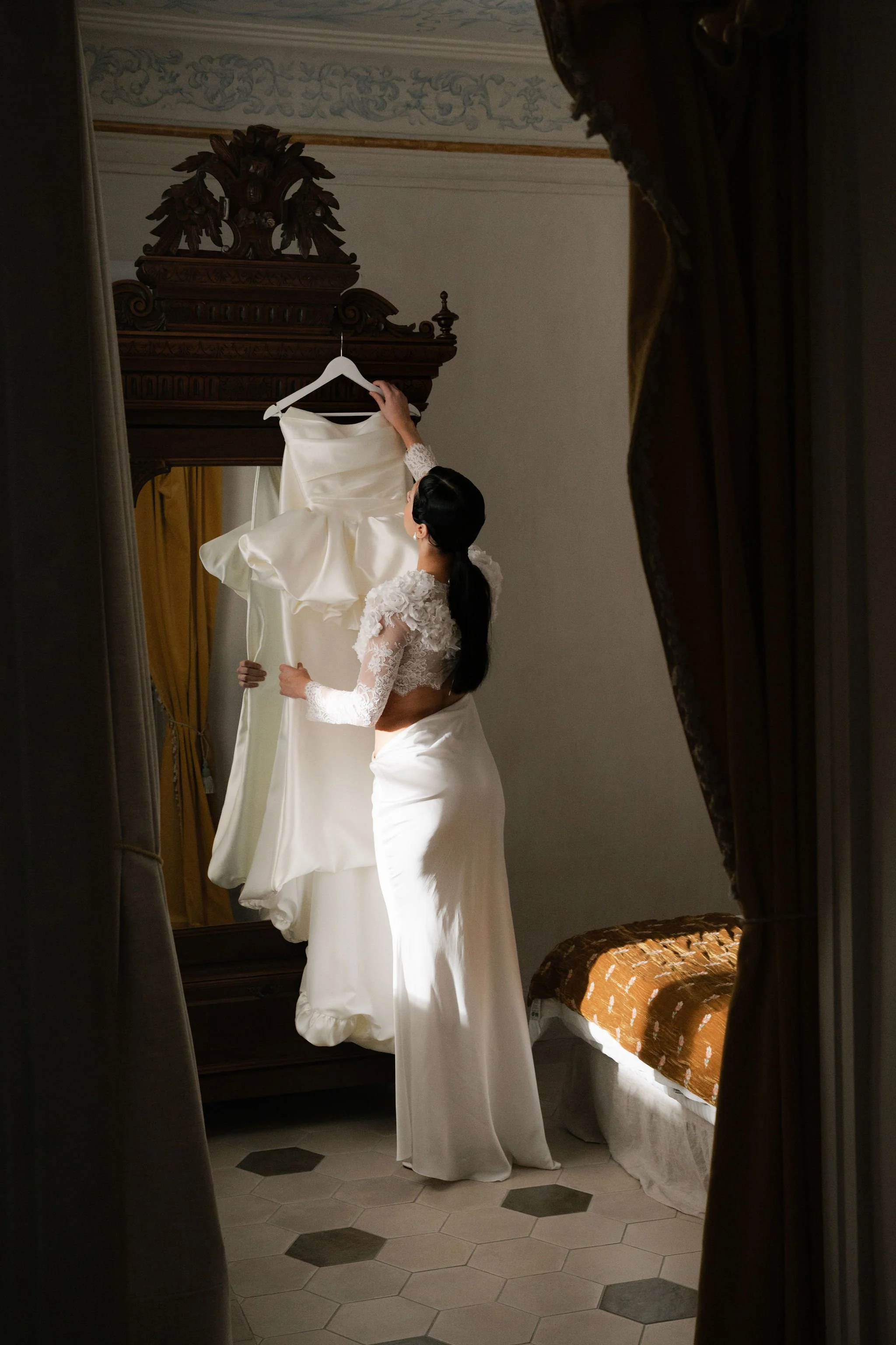A woman with dark hair in a braid, wearing a white lace dress with long sleeves, is holding up a white satin wedding dress in front of a mirror in a room with yellow curtains, a wooden ornate mirror frame, and a brown patterned sofa.