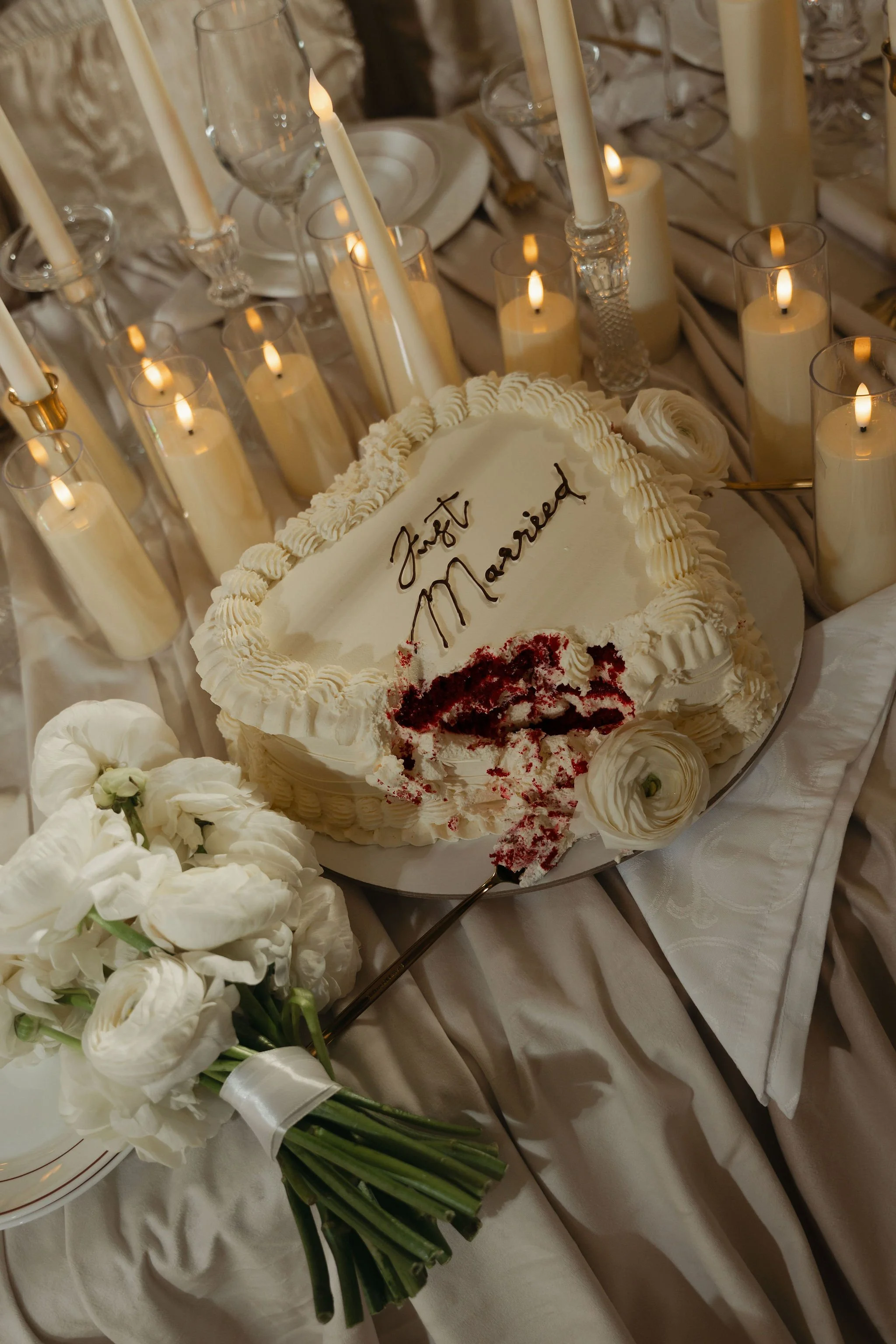 A wedding cake decorated with white cream, surrounded by lit candles and white flowers.