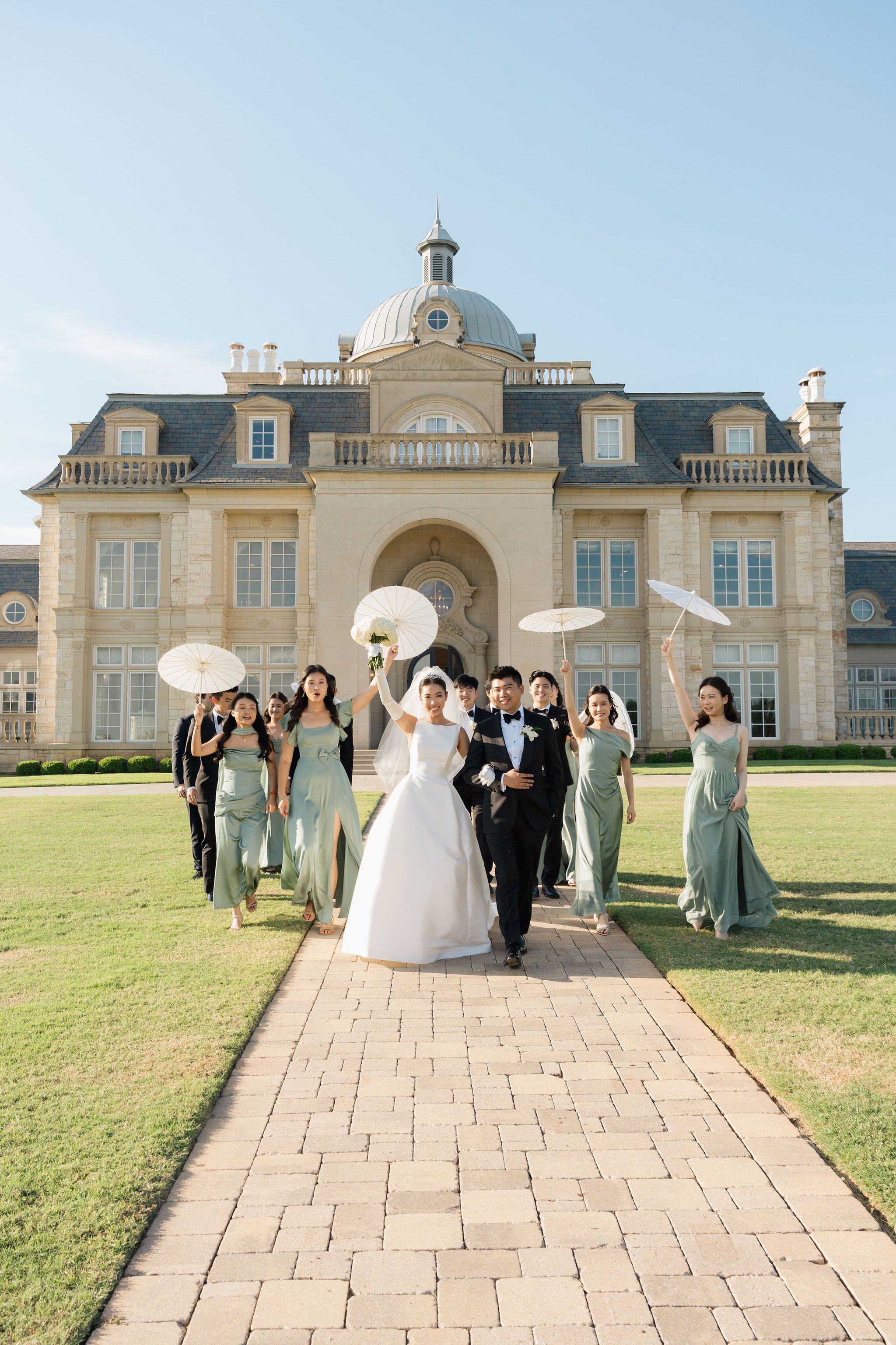 A bride and groom walk down a brick pathway while surrounded by bridesmaids and groomsmen outside a large, elegant building with a domed roof, under a clear blue sky, holding umbrellas and flower bouquets. Anna Aleksandrovna Photography