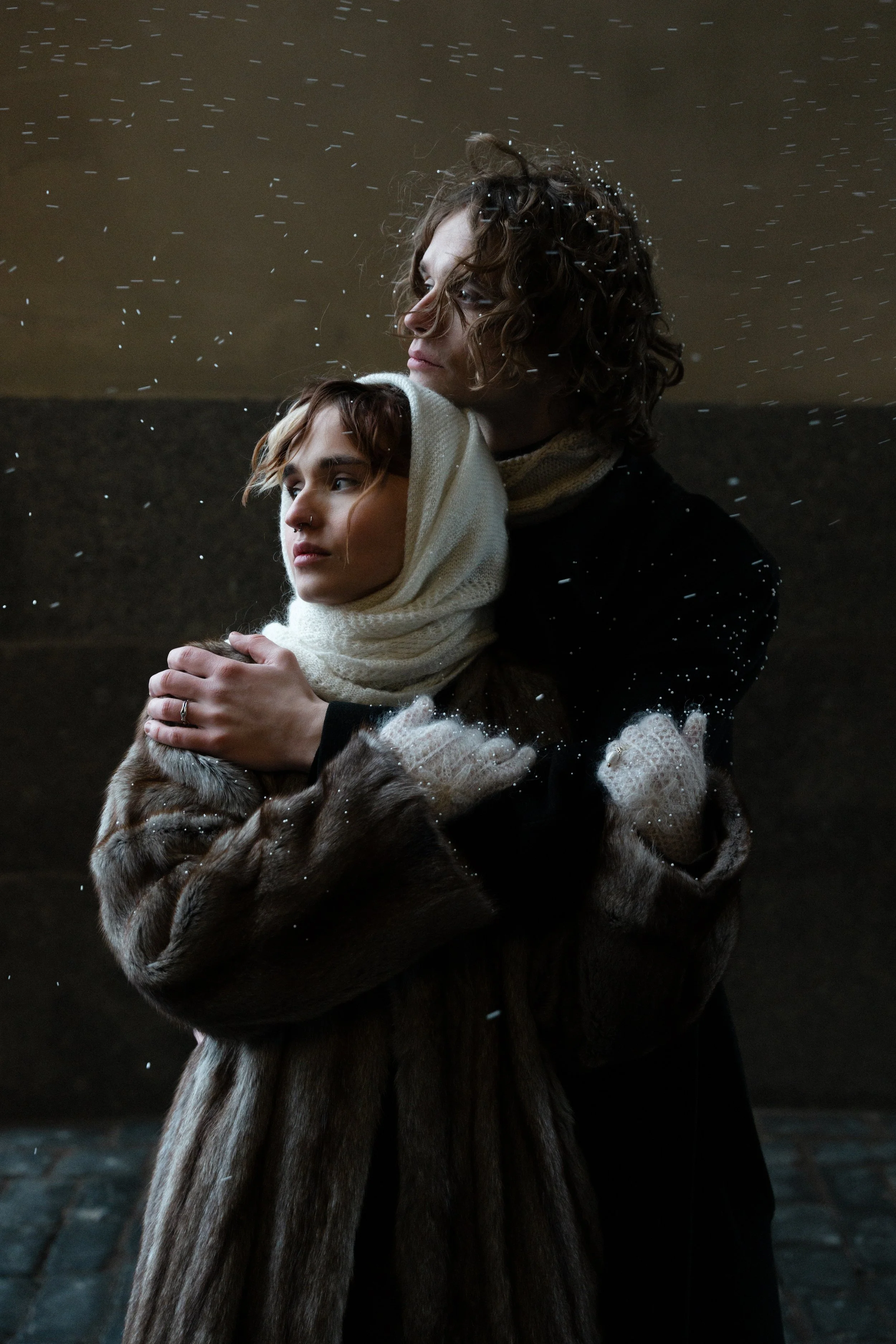 A woman wearing a fur coat and gloves is hugging another woman wrapped in a white scarf, with snow falling around them. Anna Aleksandrovna Photography