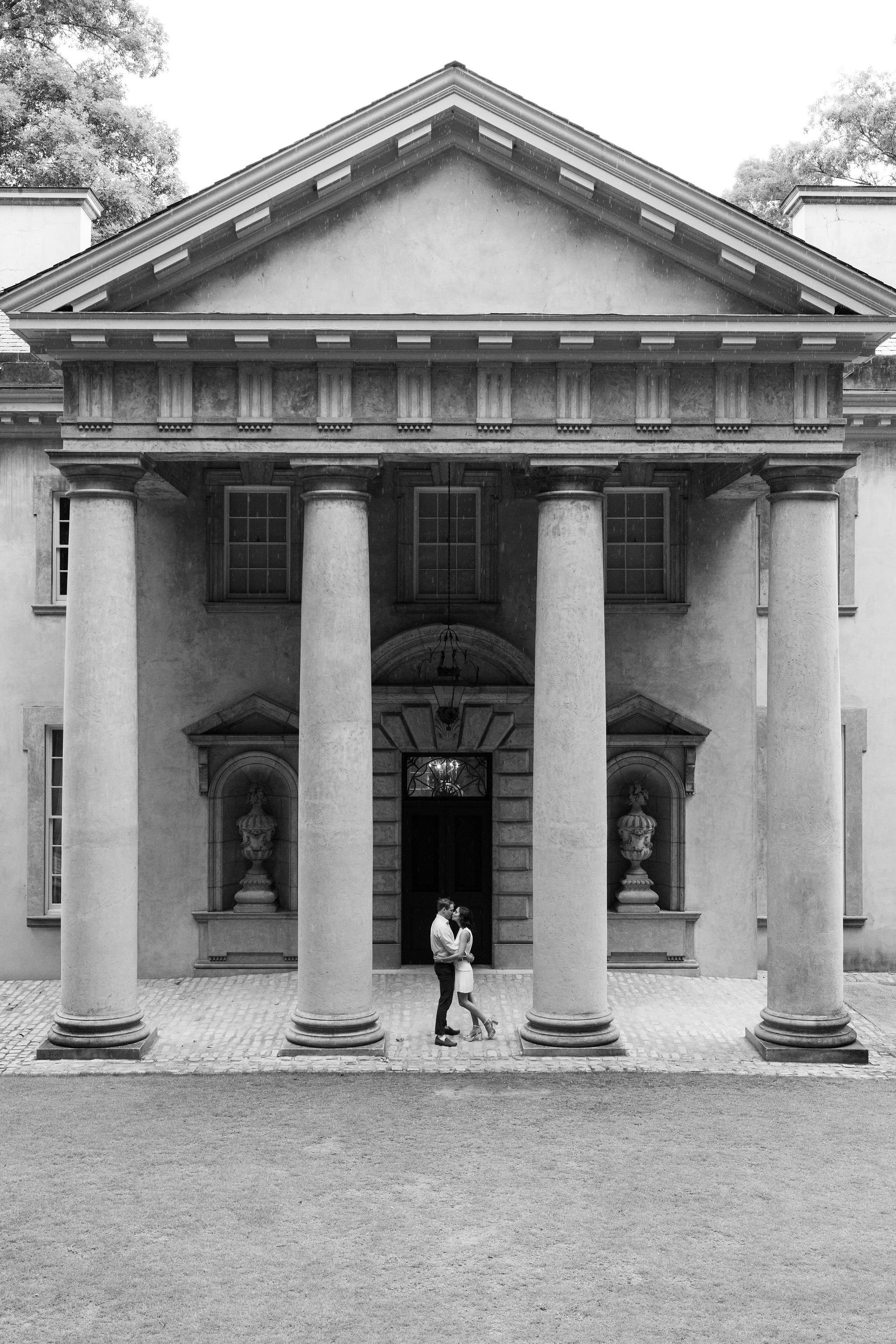 A black-and-white photo of a classical building with large columns, decorative elements, and an entrance arch. Two people, a man and a woman, are standing close together in front of the building, gazing at each other.