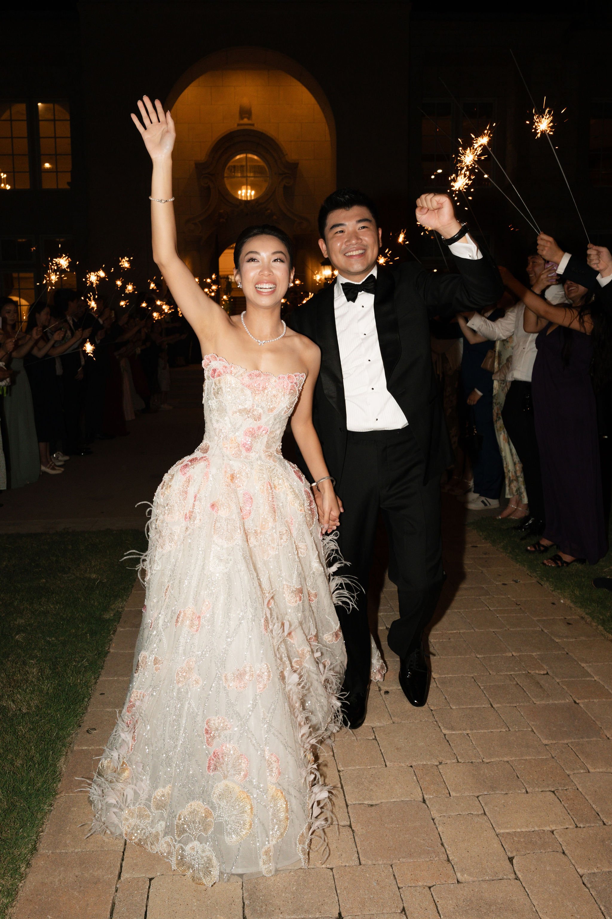 A newlywed couple celebrating at night, holding hands and smiling, surrounded by friends holding sparklers.