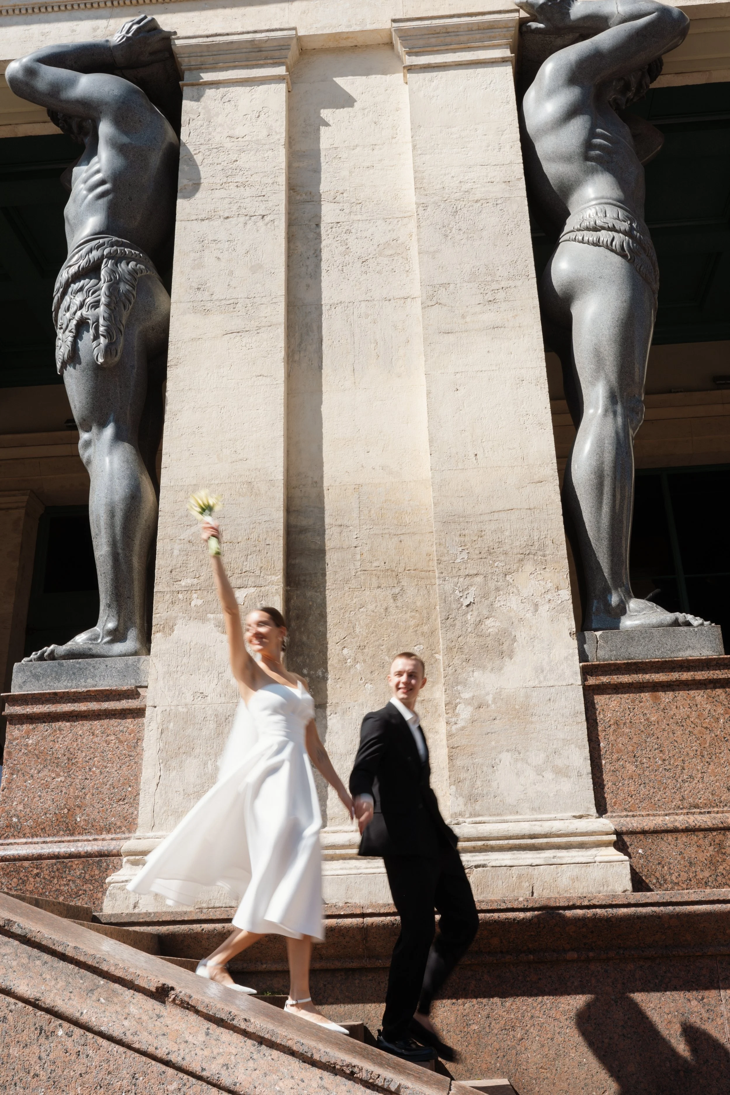 A bride and groom holding hands and walking down stone steps in front of large stone statues. The bride is wearing a white dress and holding flowers, while the groom is in a black suit. Anna Aleksandrovna Photography