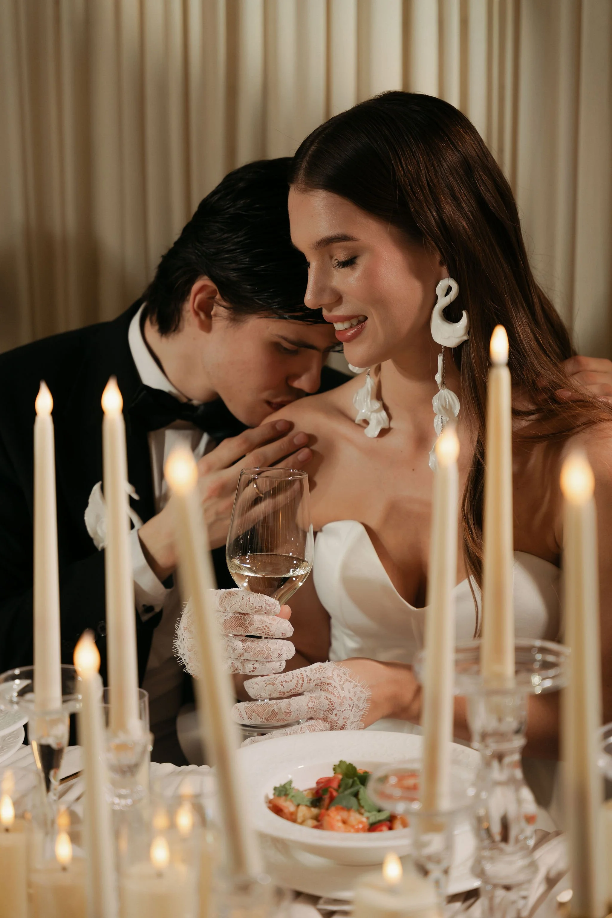 A woman in a white dress with large white earrings and long brown hair is smiling with her eyes closed, while a man in a tuxedo kisses her on the forehead. They are at an elegant dinner table with lit candles, a wine glass, and a plate of food, in fr