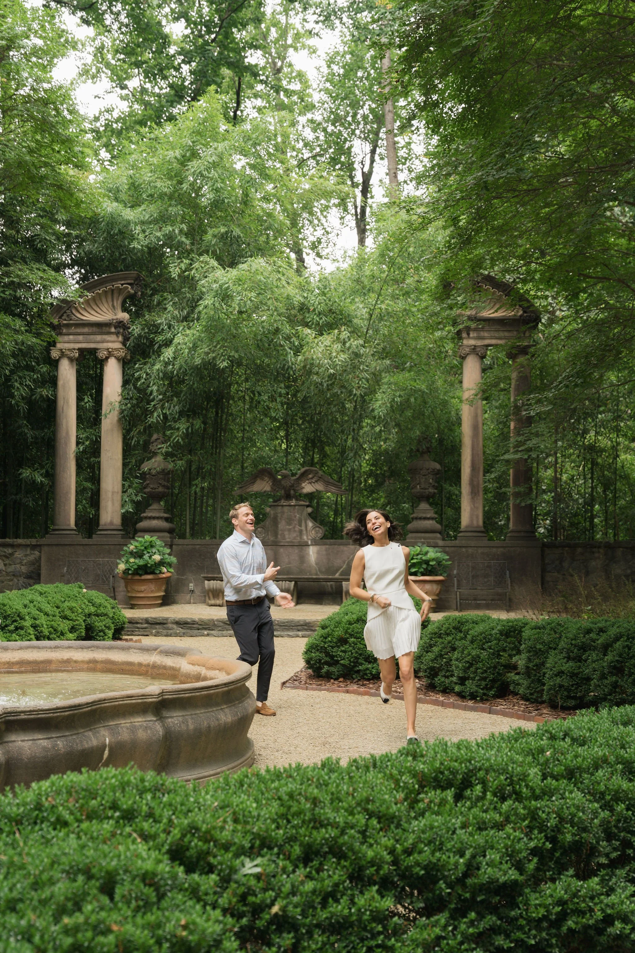 A man and a woman running and laughing in a lush garden with classical columns and green hedges.