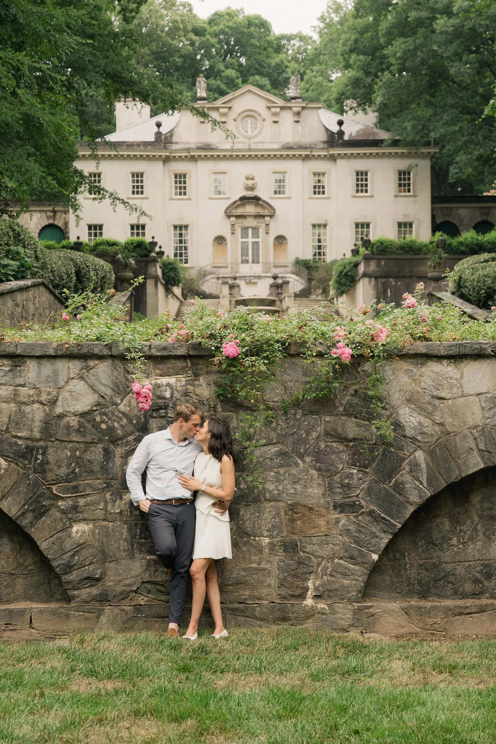 A couple kissing under a stone bridge with lush greenery and an elegant Swan House mansion in the background. Anna Aleksandrovna Photography