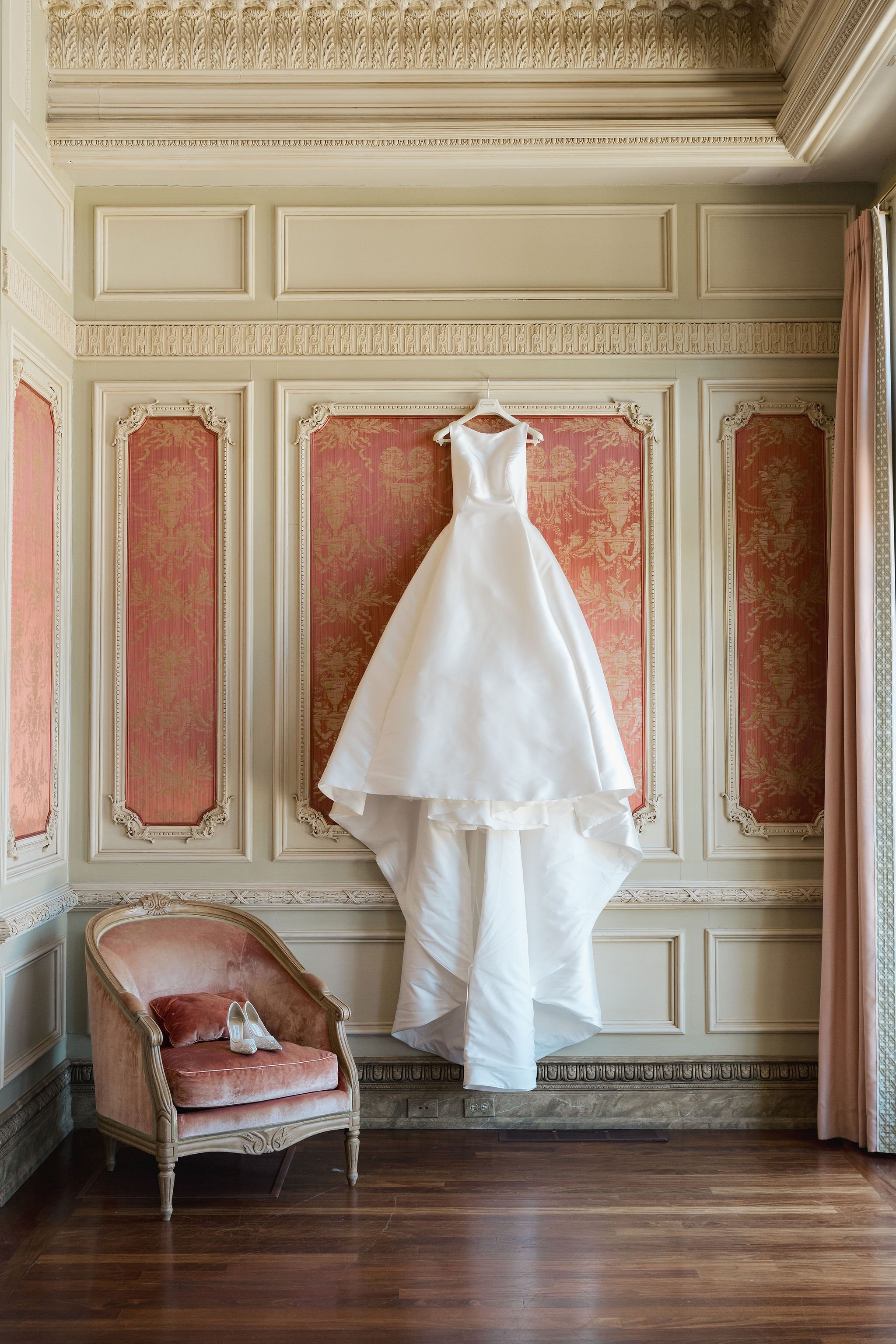 A white wedding dress hanging on a hook in front of a pink and cream ornate wall, next to a pink velvet chair with white shoes on it, in an elegant room with classical decor.