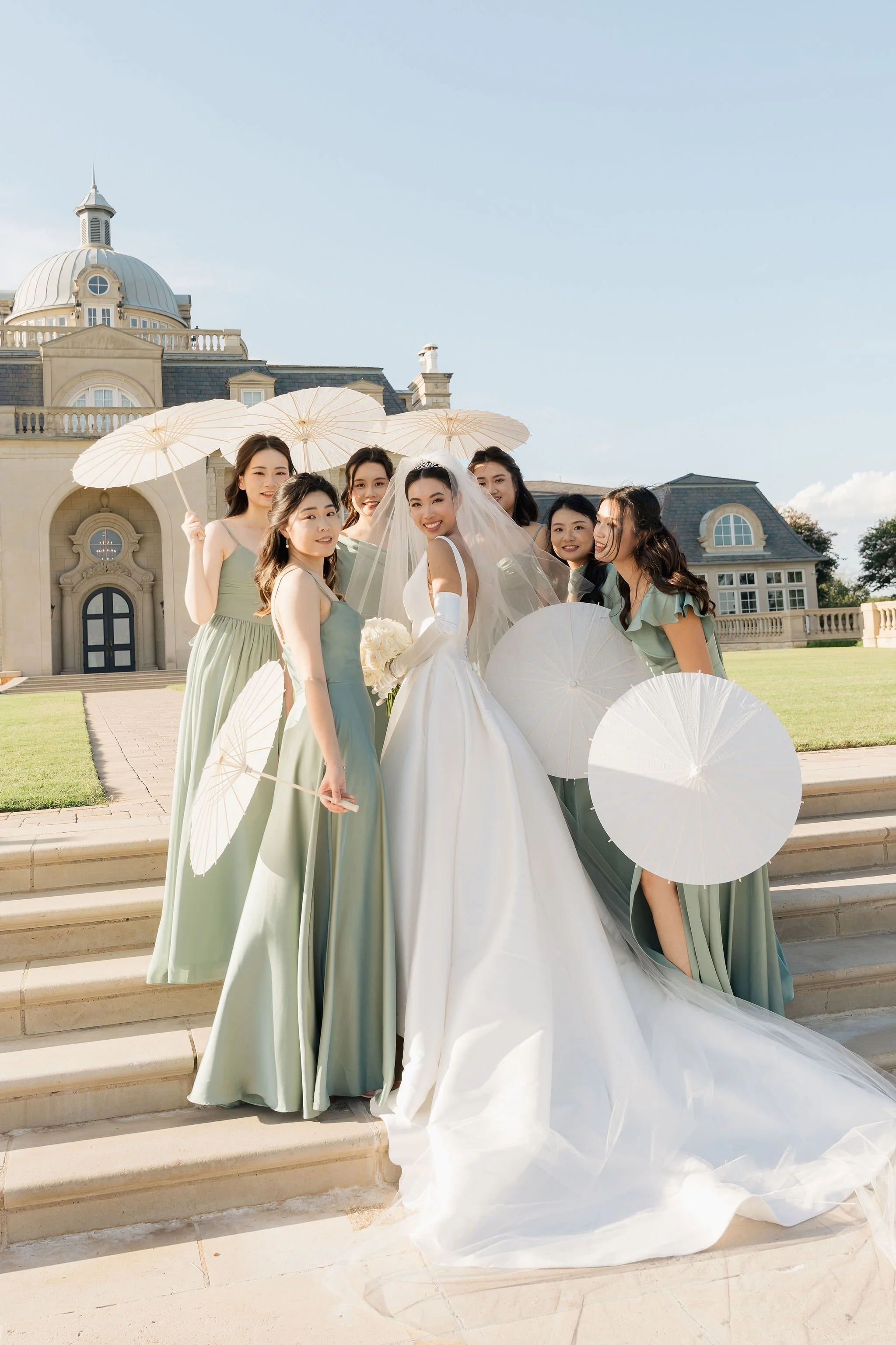bride in a white wedding gown with a veil, and bridesmaids in light green dresses, standing on the stairs in front of The Olana in Dallas TX. They are holding white parasols and smiling for a photo. Anna Aleksandrovna Photography