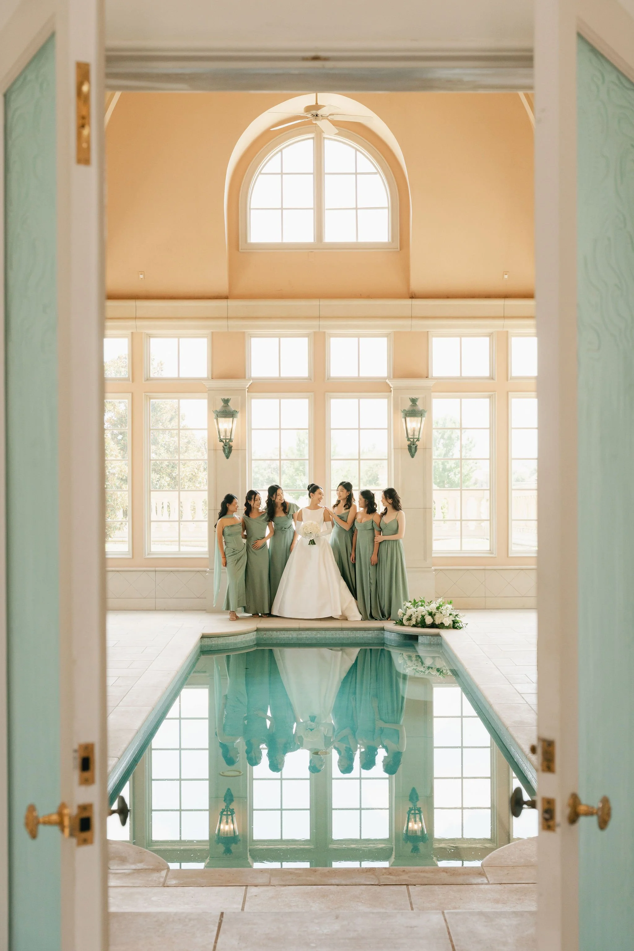 A bride and six bridesmaids standing in front of large windows and a pond, seen through an open doorway with a reflecting pool in the foreground.