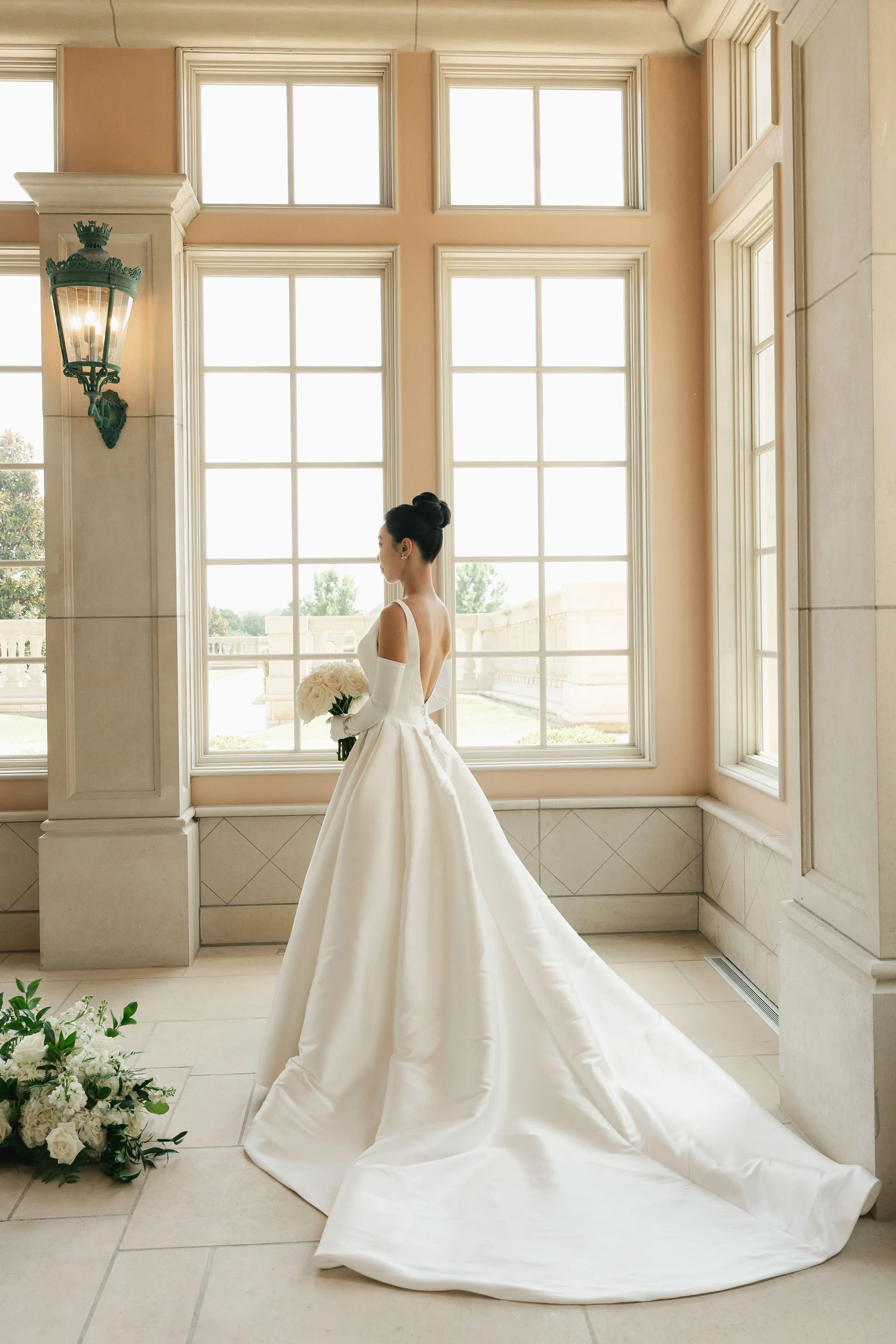 A woman dressed in a white wedding gown holding a bouquet of white roses, standing in front of large windows in an elegant room with cream-colored walls and decorative moldings.