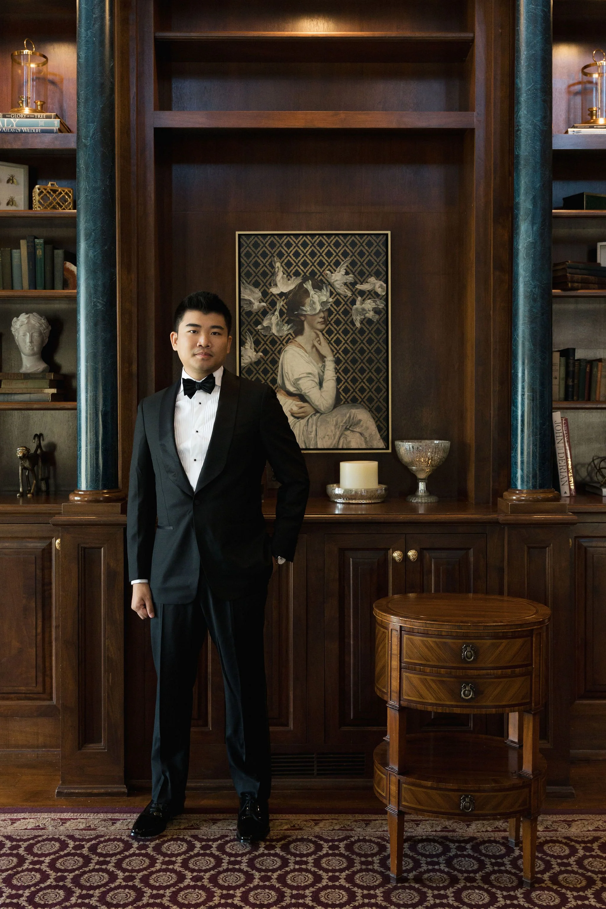 Young man in black tuxedo standing in front of a wooden bookshelf in a formal setting, with a painting of a woman surrounded by swans on the wall behind him.