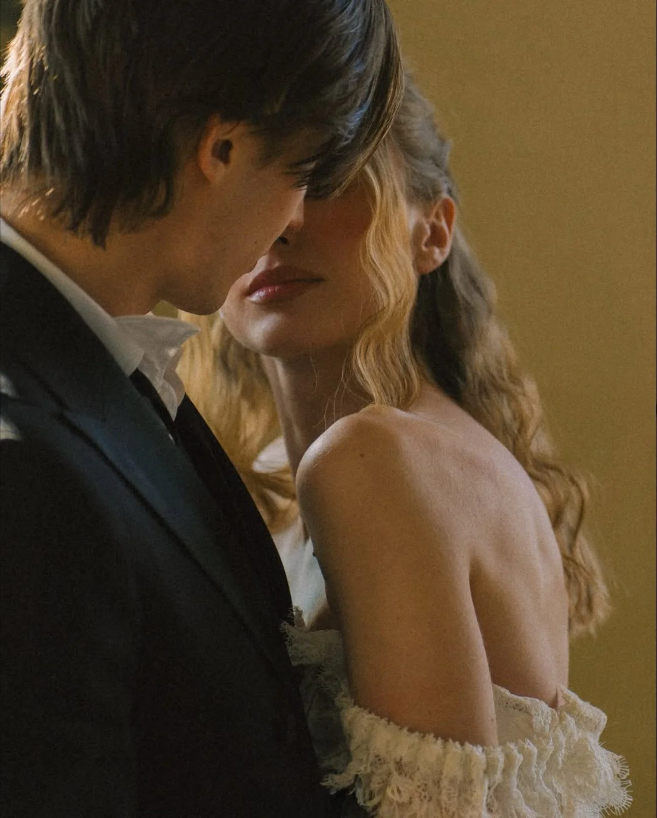 A close-up of a man in a black suit and a woman with long, curly hair, with their forehead and nose touching in an intimate moment.