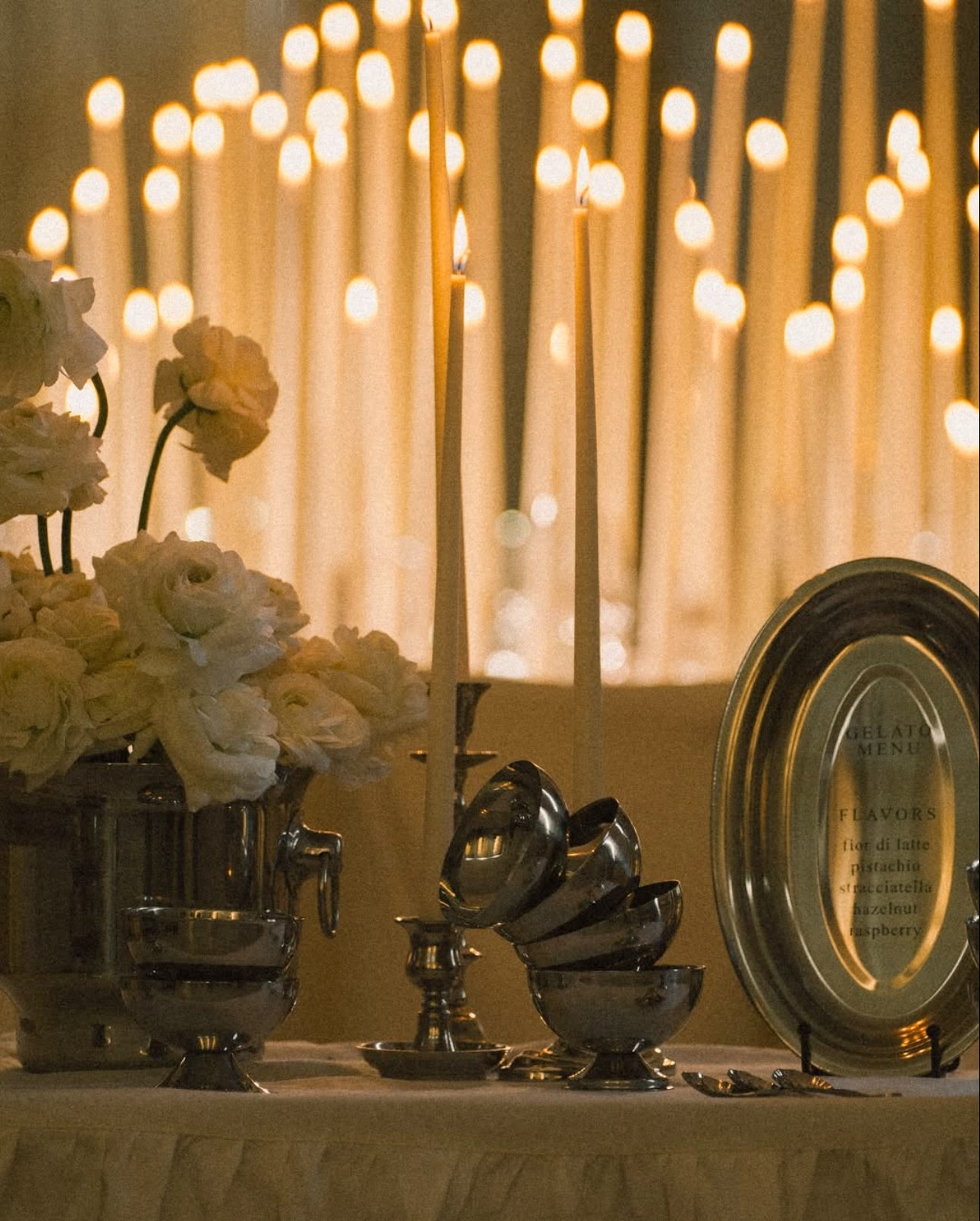 Silver trays and bowls with a bouquet of white roses, lit candles, a menu plate listing flavors, and a blurred background of hanging string lights.