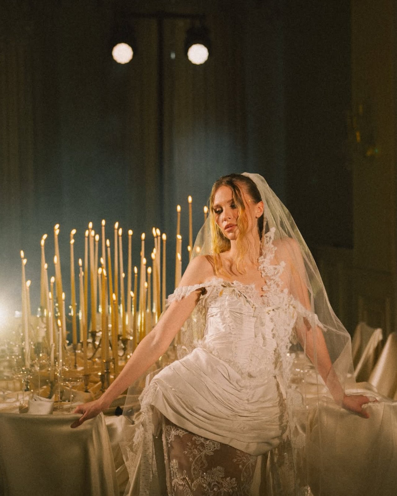 A woman in a wedding dress sitting at a table with lit candles in the background. Anna Aleksandrovna Photography