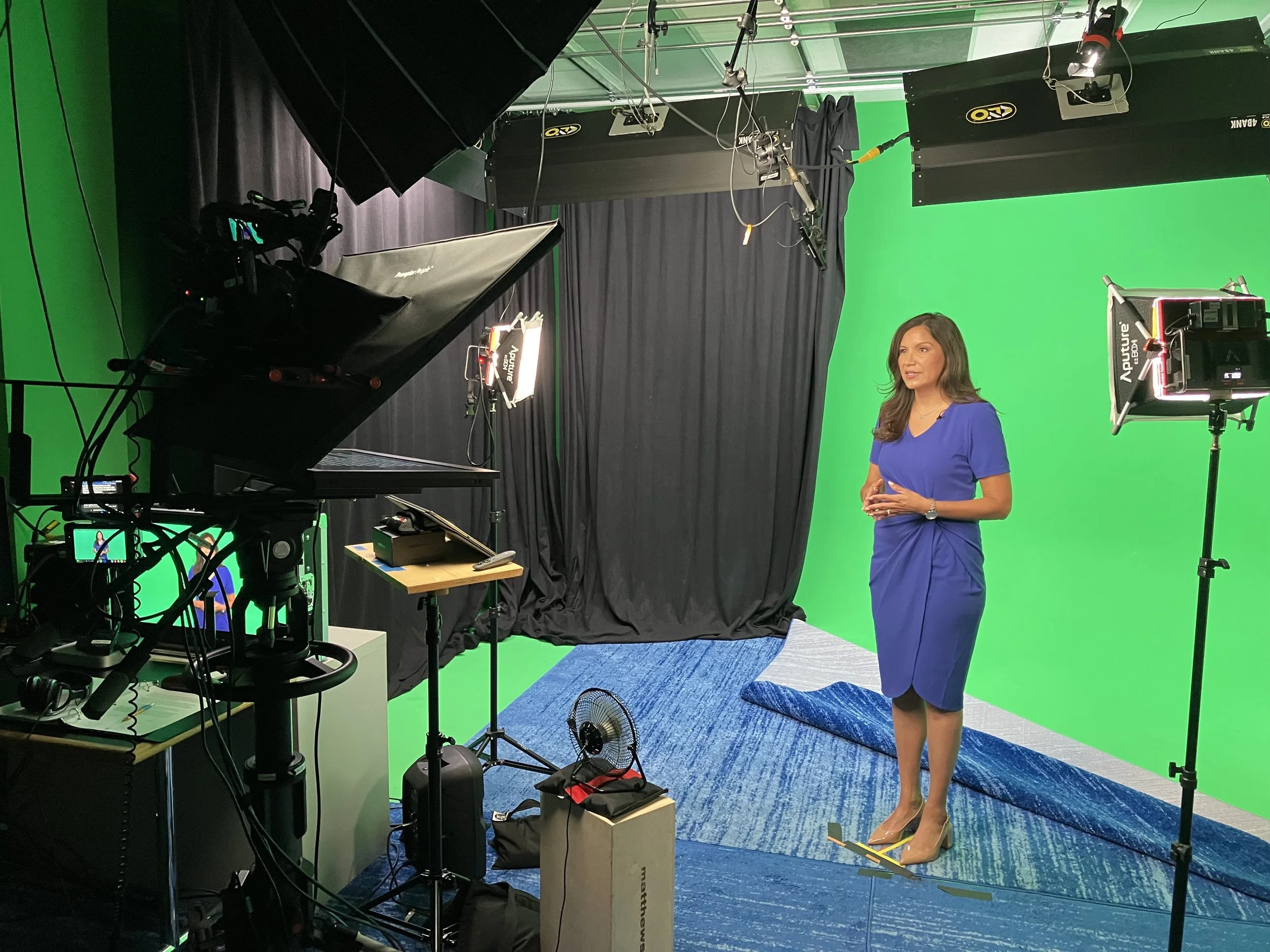 A woman in a blue dress standing in front of a green screen in a professional recording studio, with video and camera equipment around her.