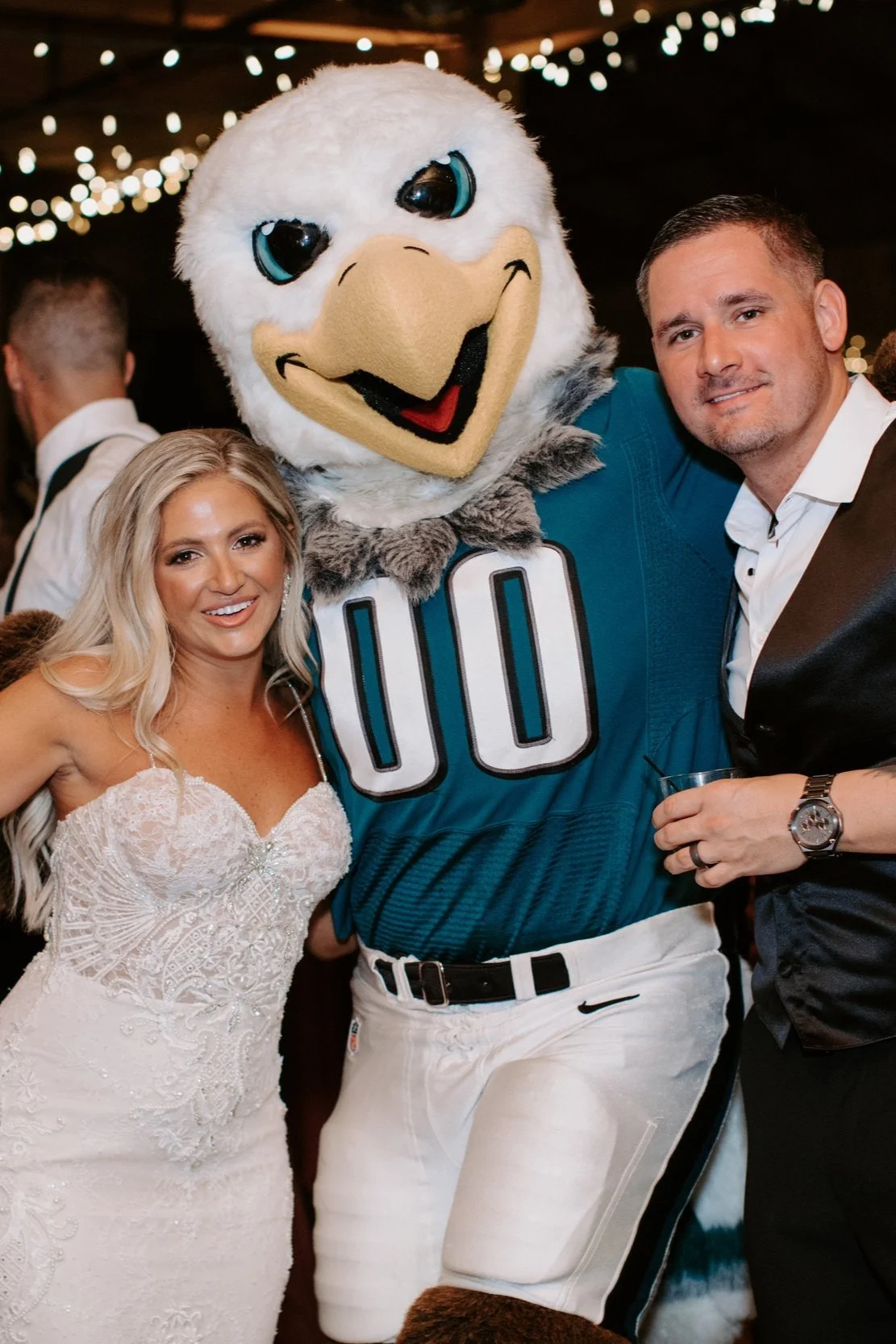 A woman in a white wedding dress, a person in a bird mascot costume wearing a sports jersey with the number 00, and a man in a black tuxedo posing together at a celebration or party, with string lights in the background.