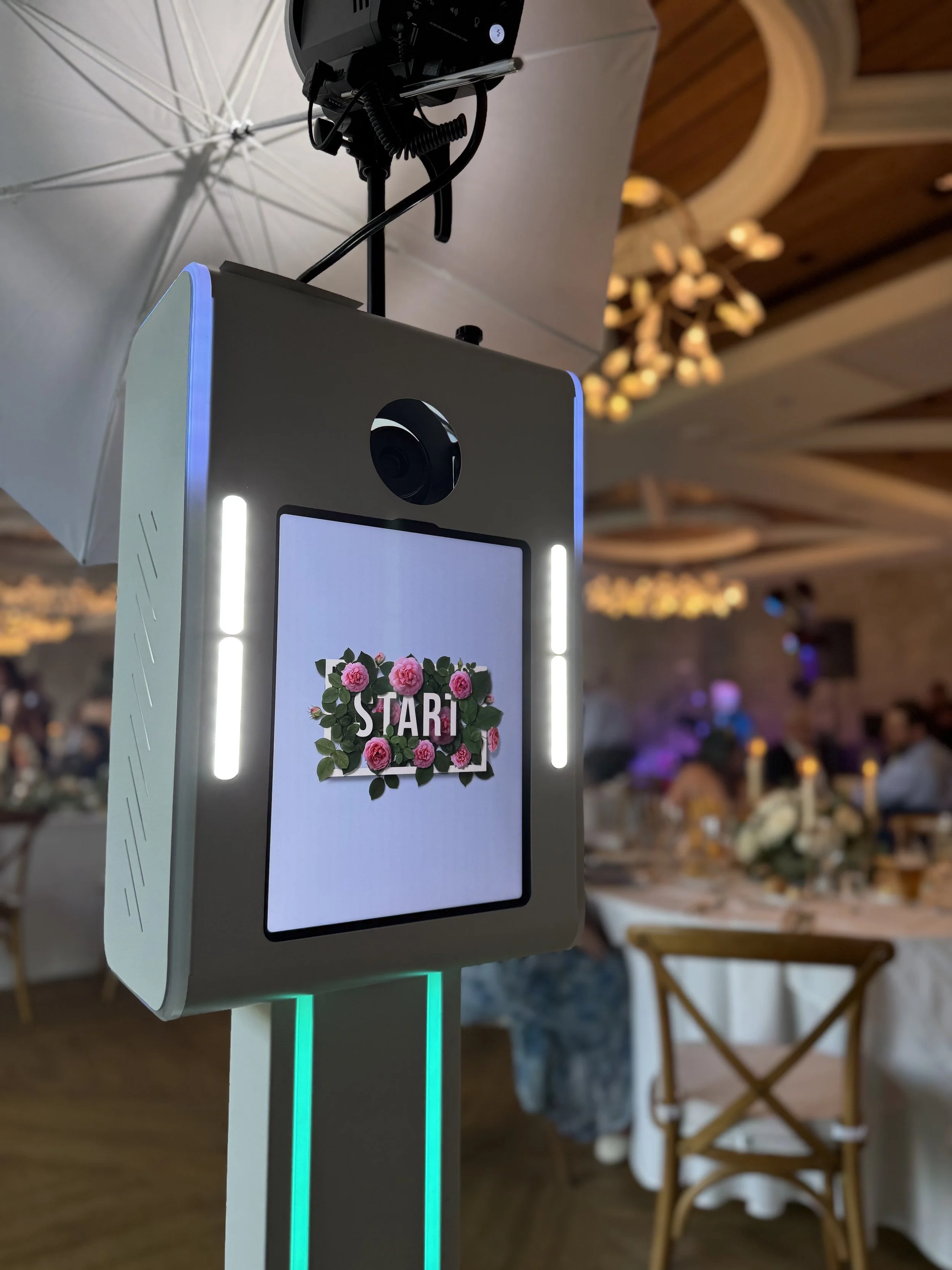 A digital wedding sign with a black-and-white photo of a couple, and flowing text that says "Welcome to the wedding of Angelina & Michael, June 10th, 2023". The sign is illuminated and set against a lush green leafy background with a glowing "Mr & Mrs" sign.