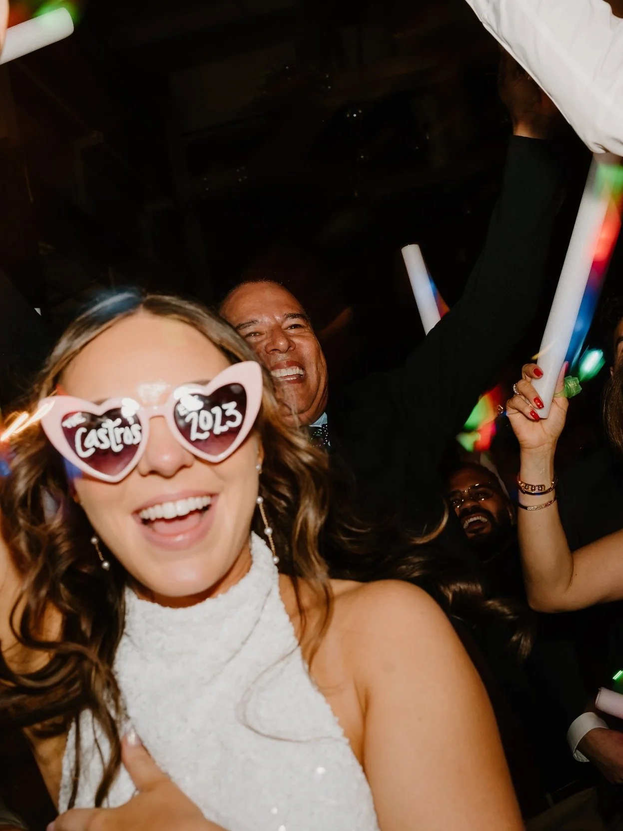 Celebrating a beautiful wedding in 2023 with smiling bride wearing heart-shaped glasses and holding glowing sticks at a party.