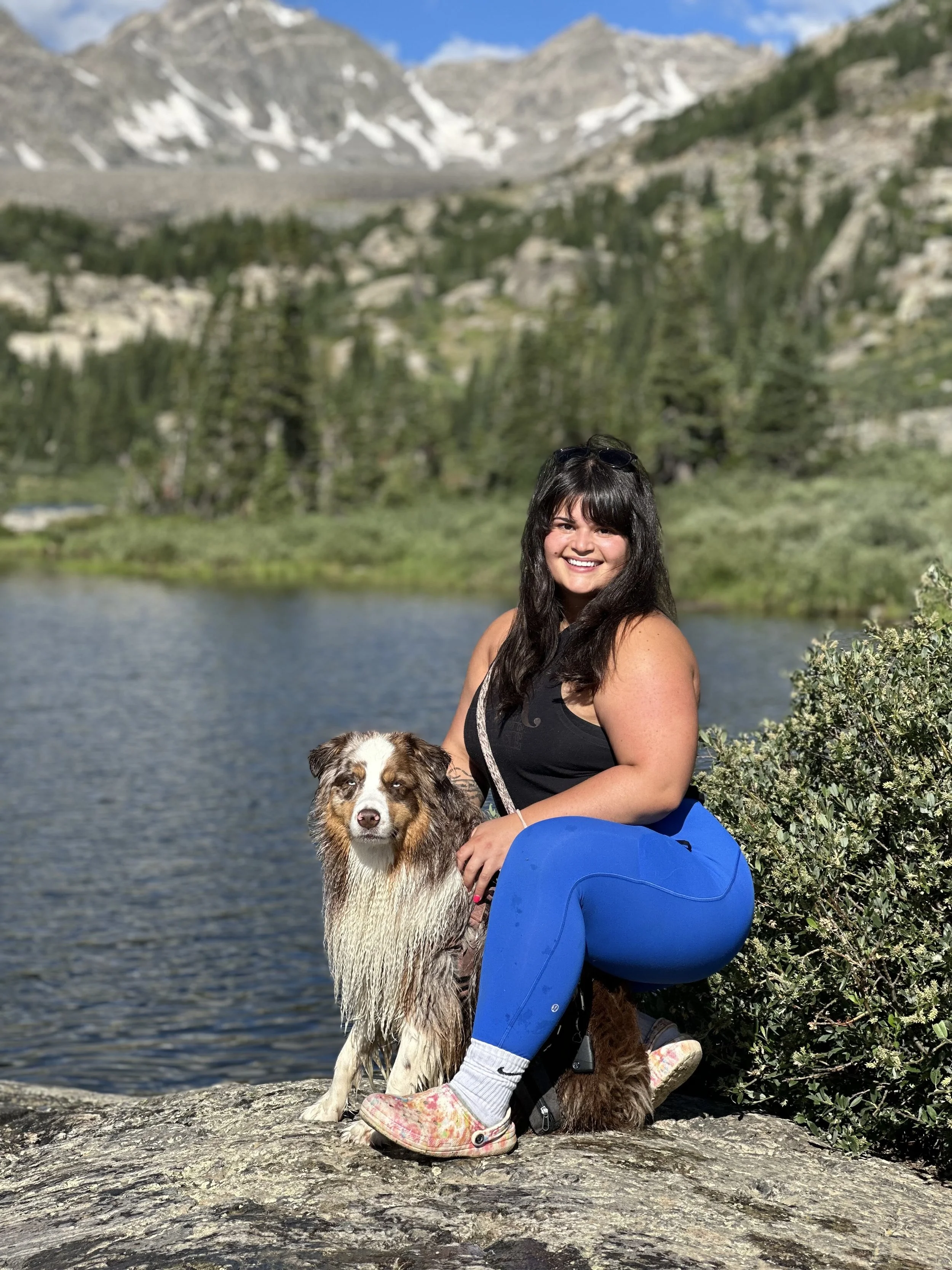 A picture of Andrea Cerreta, Golden PT & Pilates receptionist, smiling and sitting on a rock by a lake, holding a dog with a tri-color coat, both surrounded by mountains and trees on a sunny day.