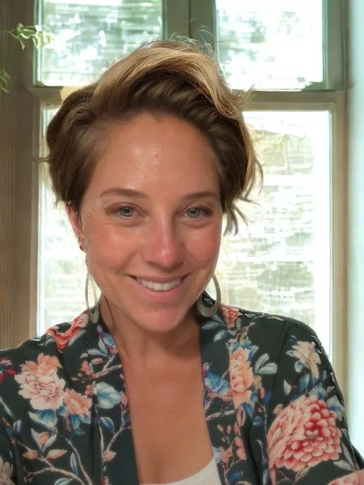 A picture of Jo Pennock, Golden PT & Pilates receptionist, with short, styled brown hair smiling inside a room with large windows and greenery outside.