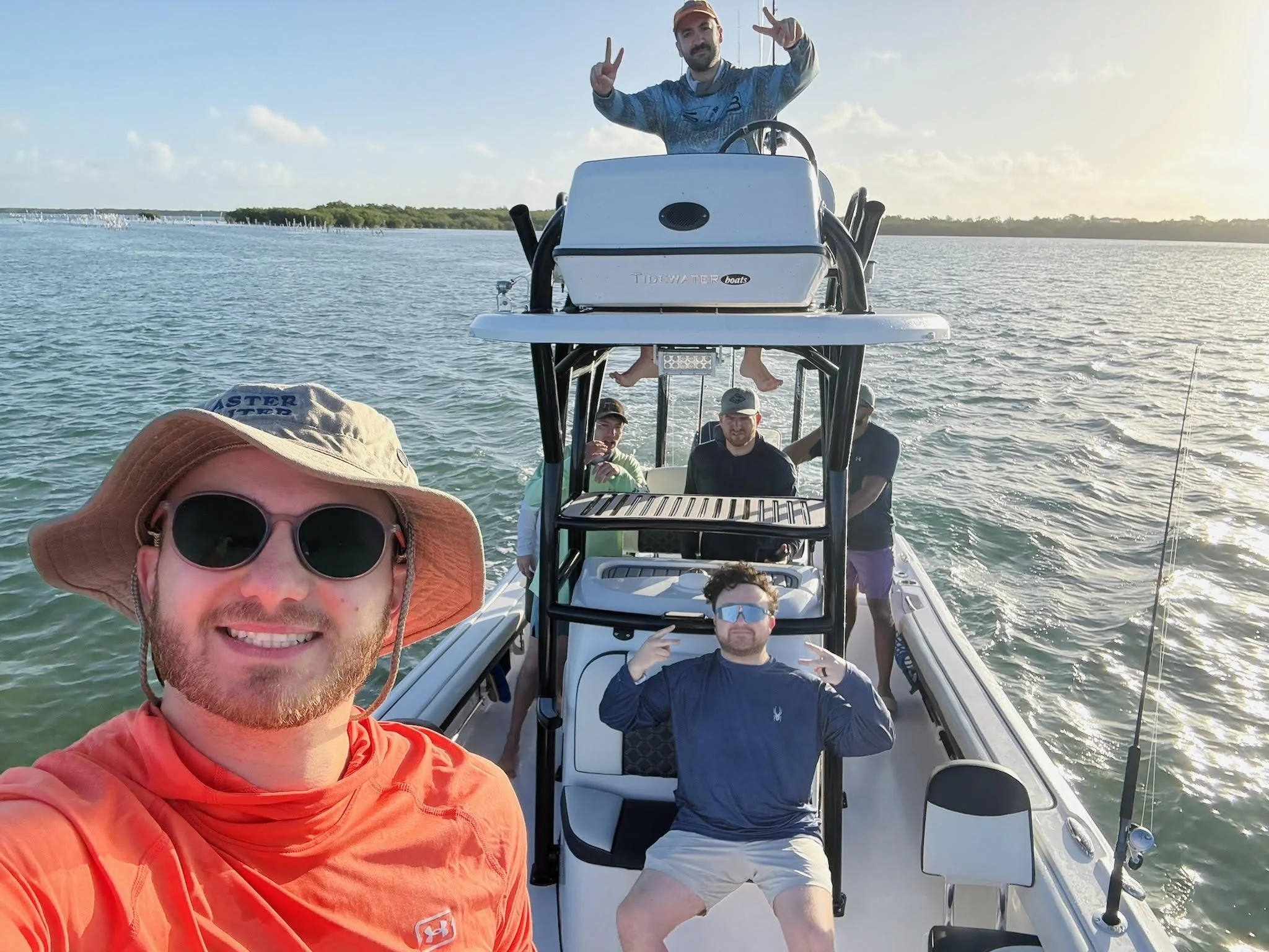 A group of people on a boat enjoying a day of fishing and boating on a body of water, with some people smiling and one person making a peace sign, in the late afternoon sunlight.