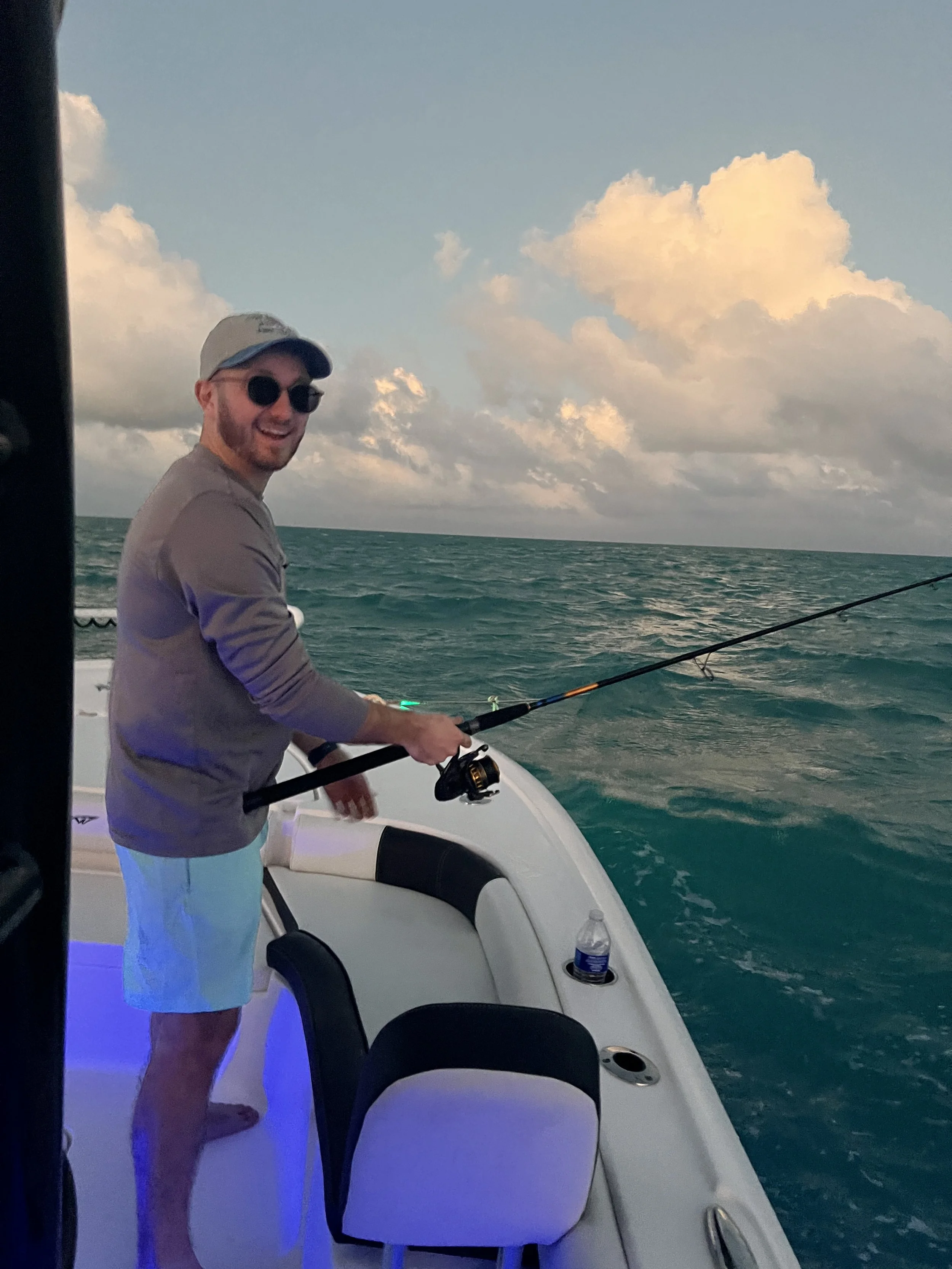A man is fishing from a boat on open water with a cloudy sky in the background. He is wearing sunglasses, a hat, a long-sleeved shirt, and shorts.
